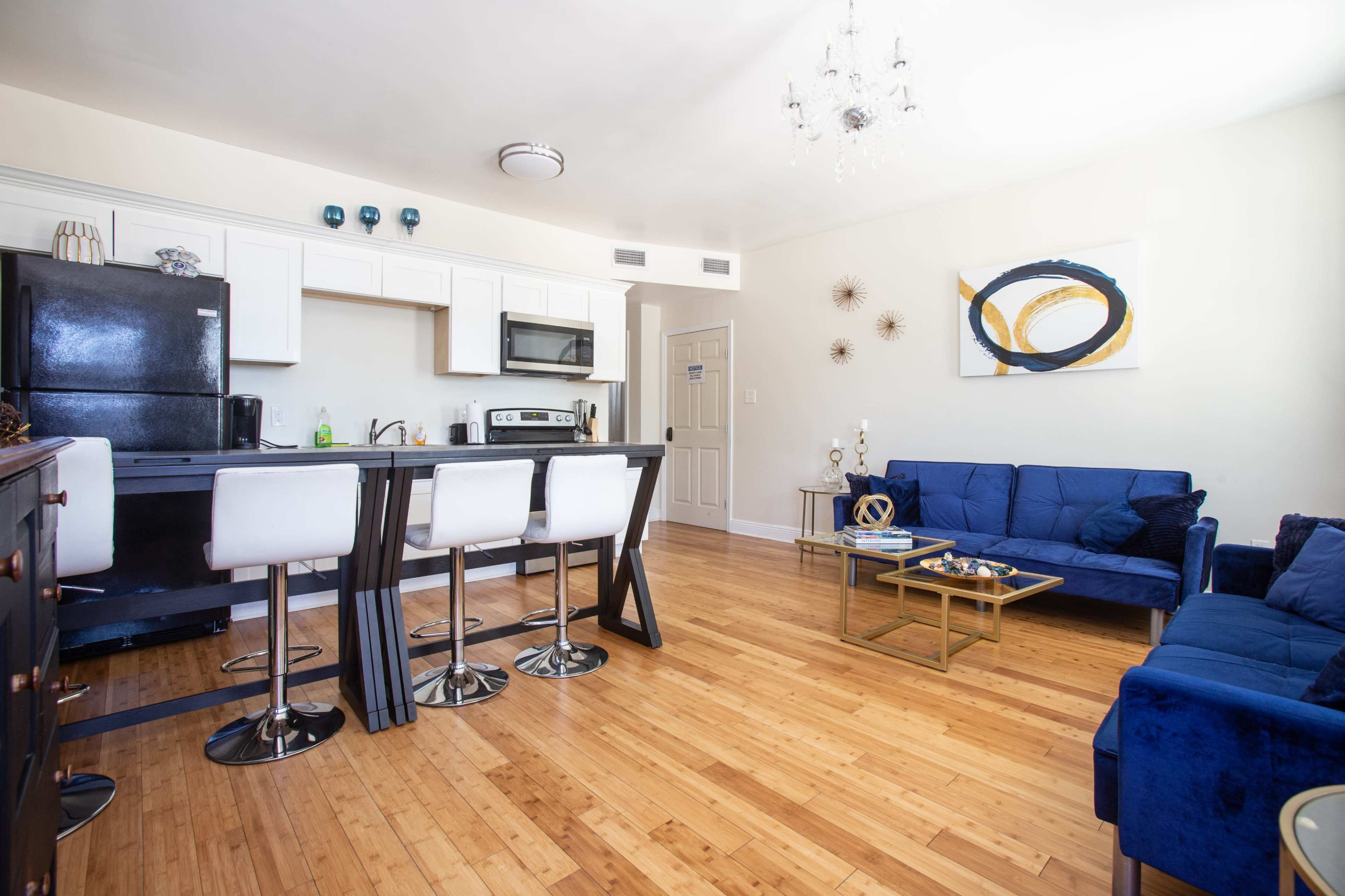 A modern living space featuring a kitchen bar with white stools, a dark refrigerator, and a seating area with blue upholstery and a glass coffee table.