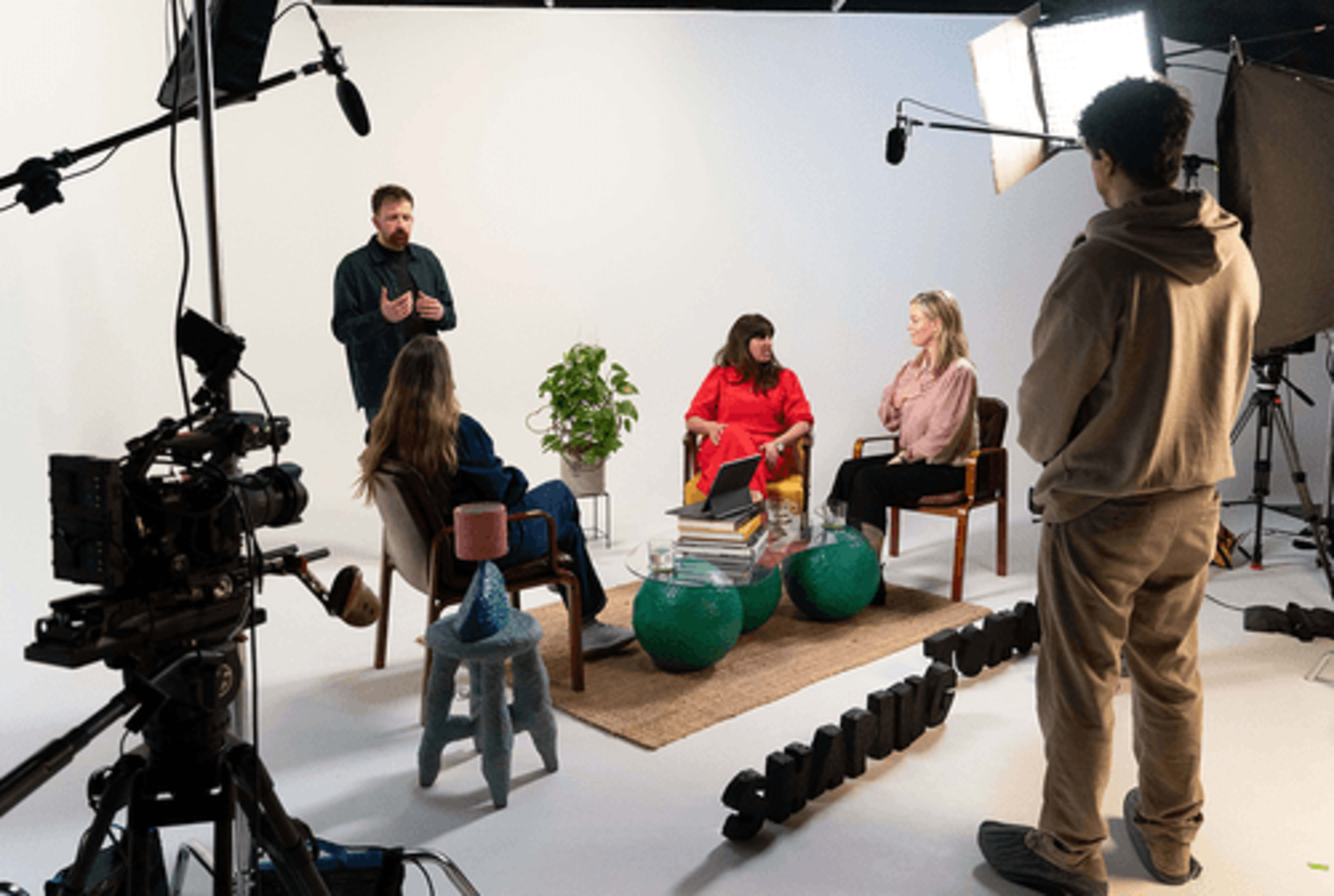 A group of four people is engaged in discussion on a set with a minimalist background, surrounded by cameras and lighting equipment.