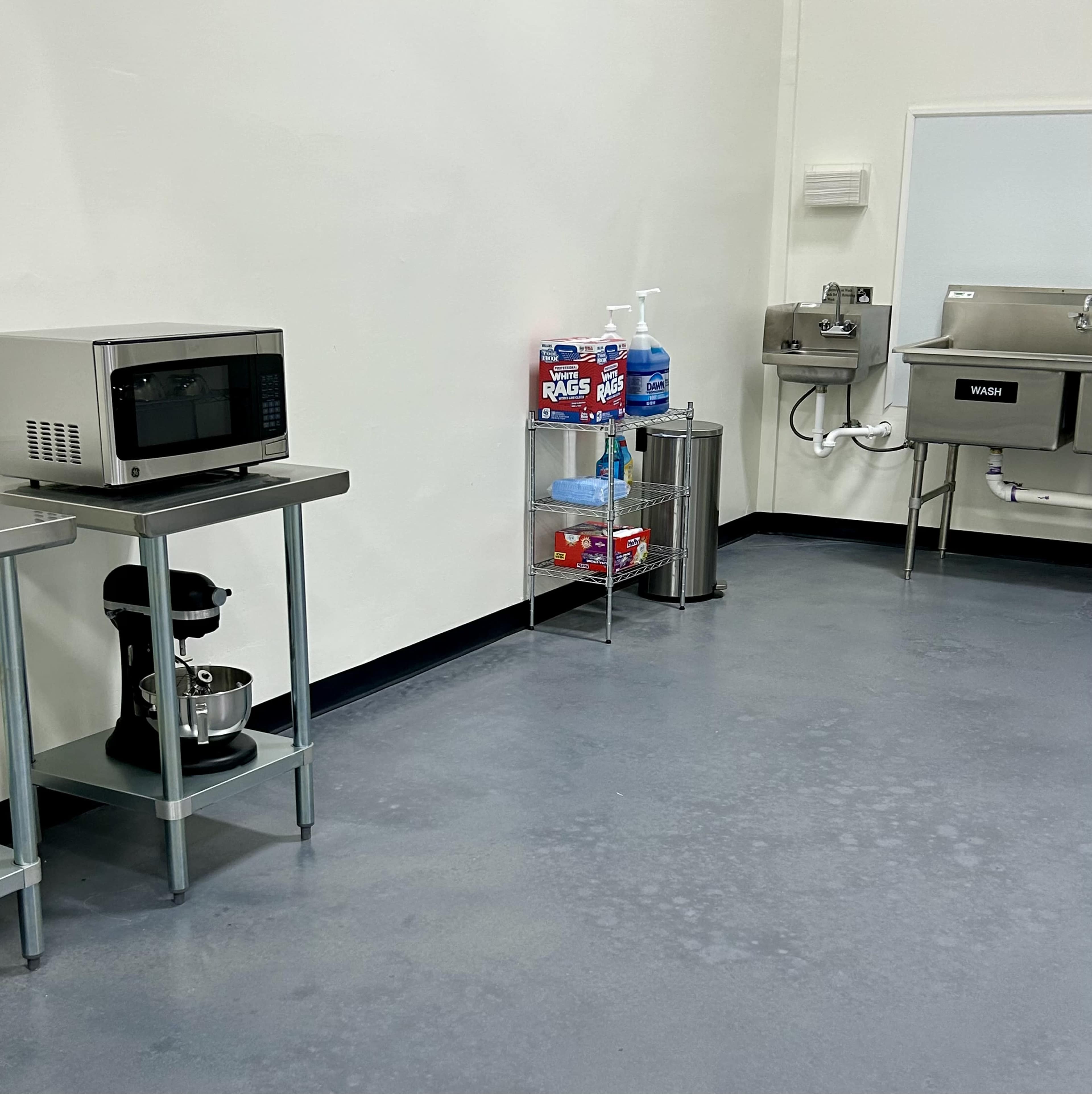The image shows a clean kitchen space featuring a microwave, a stand mixer on a table, a handwashing sink, and cleaning supplies on a shelf.
