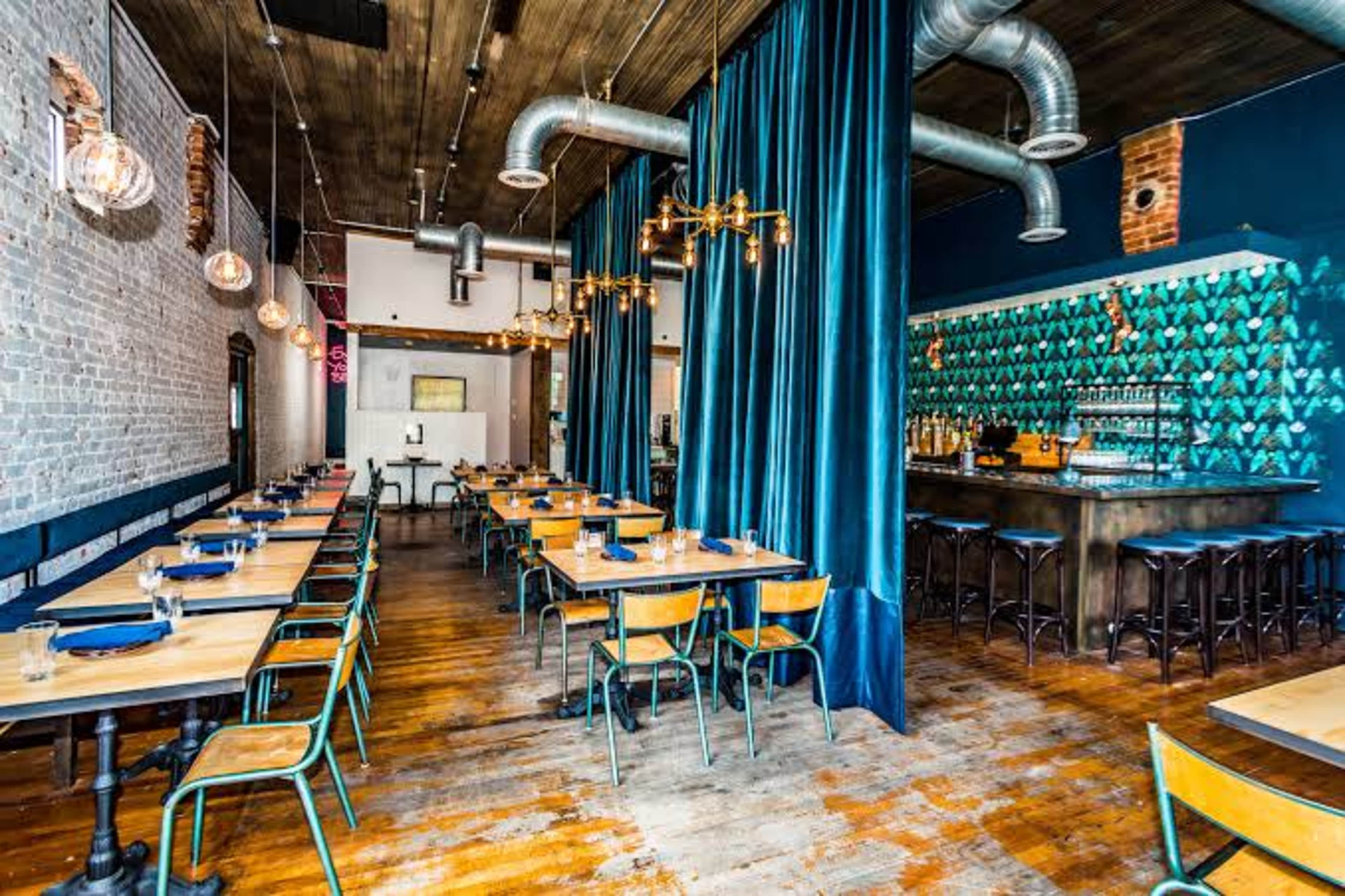 The image shows a modern restaurant interior featuring wooden floors, blue velvet curtains, and a bar with intricate tile work.