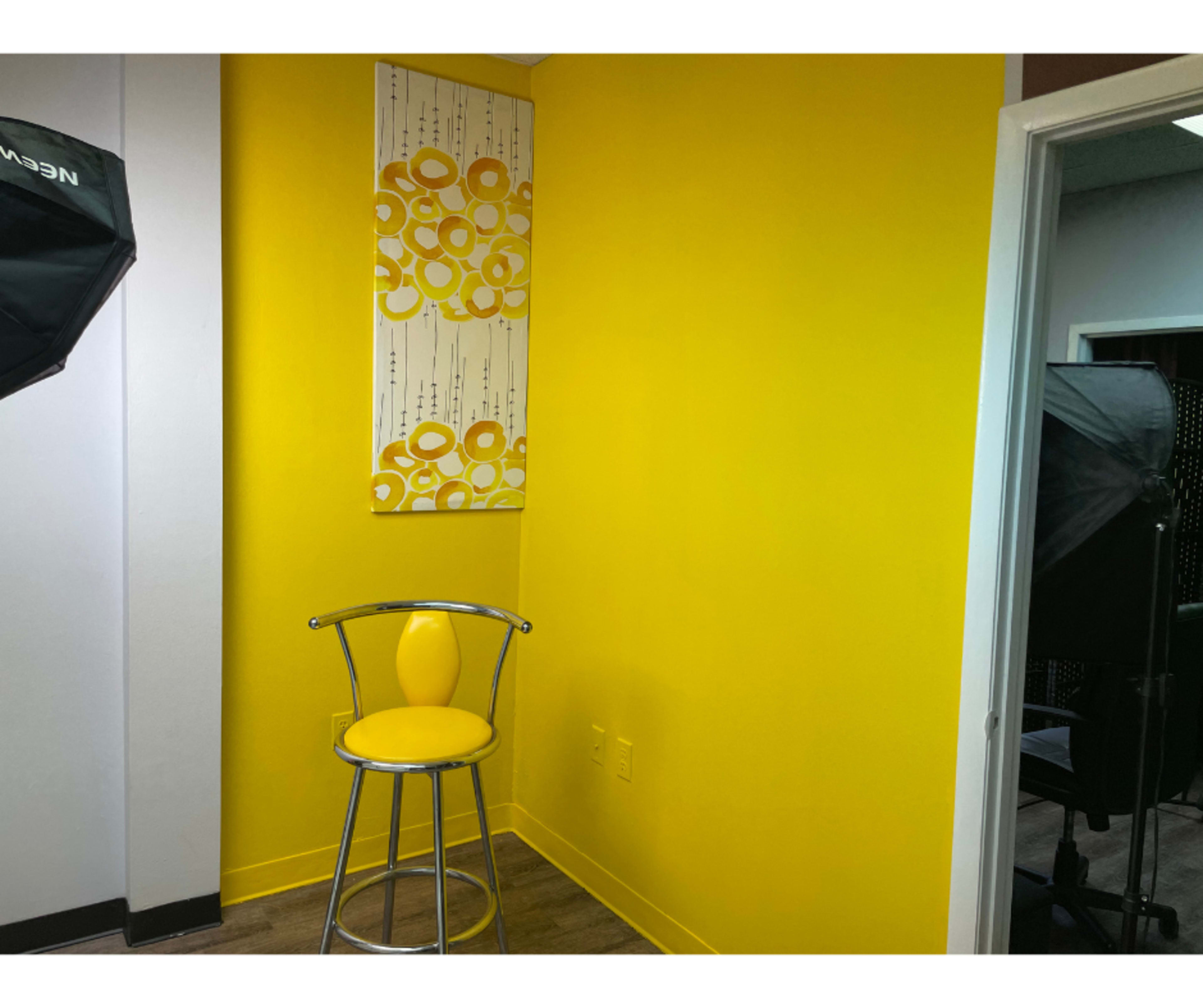 A yellow-walled corner features a silver stool with a matching seat and an abstract artwork above it.