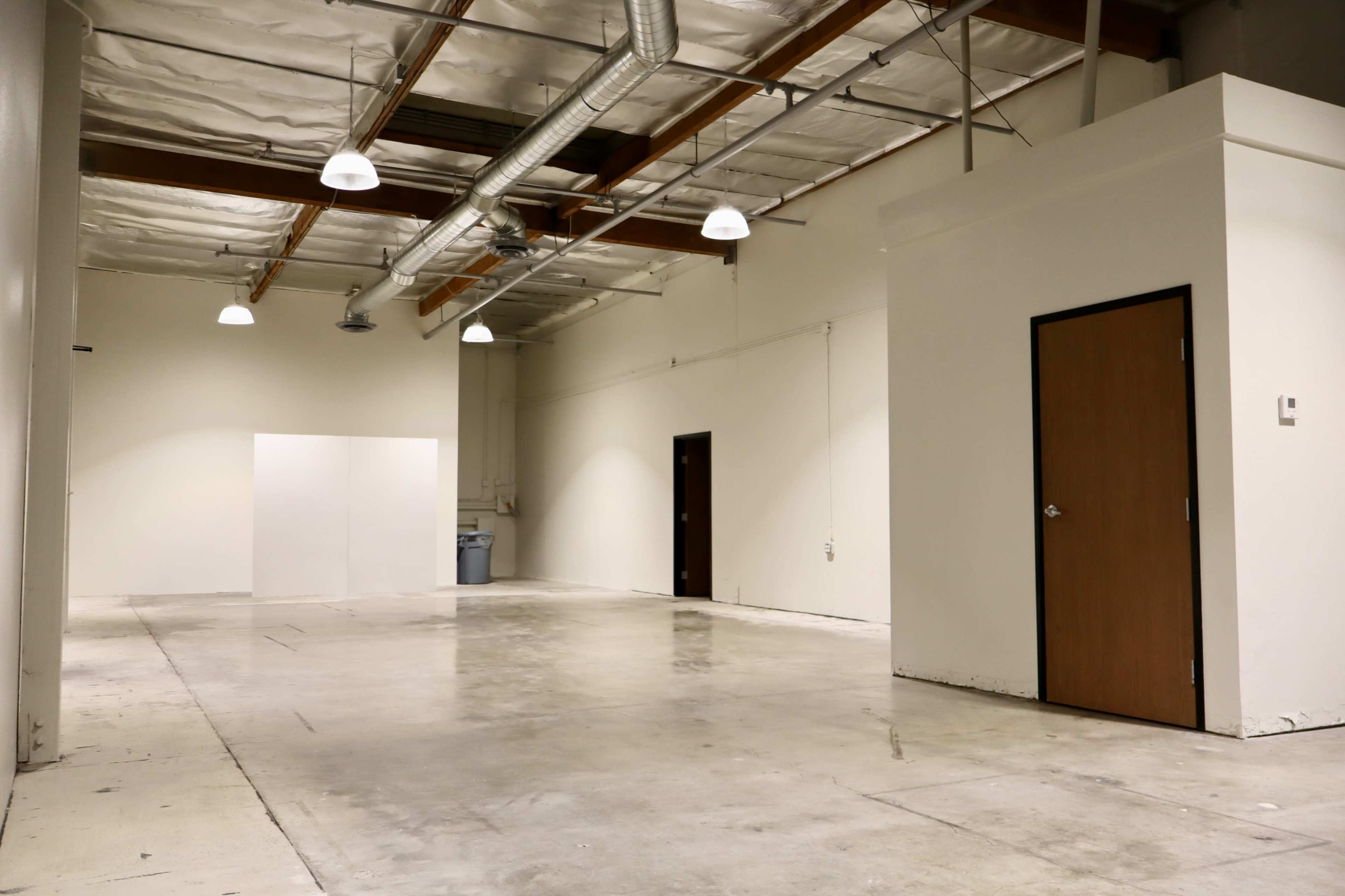 The image shows a large, empty industrial space with concrete flooring, white walls, and a single door on the right side.
