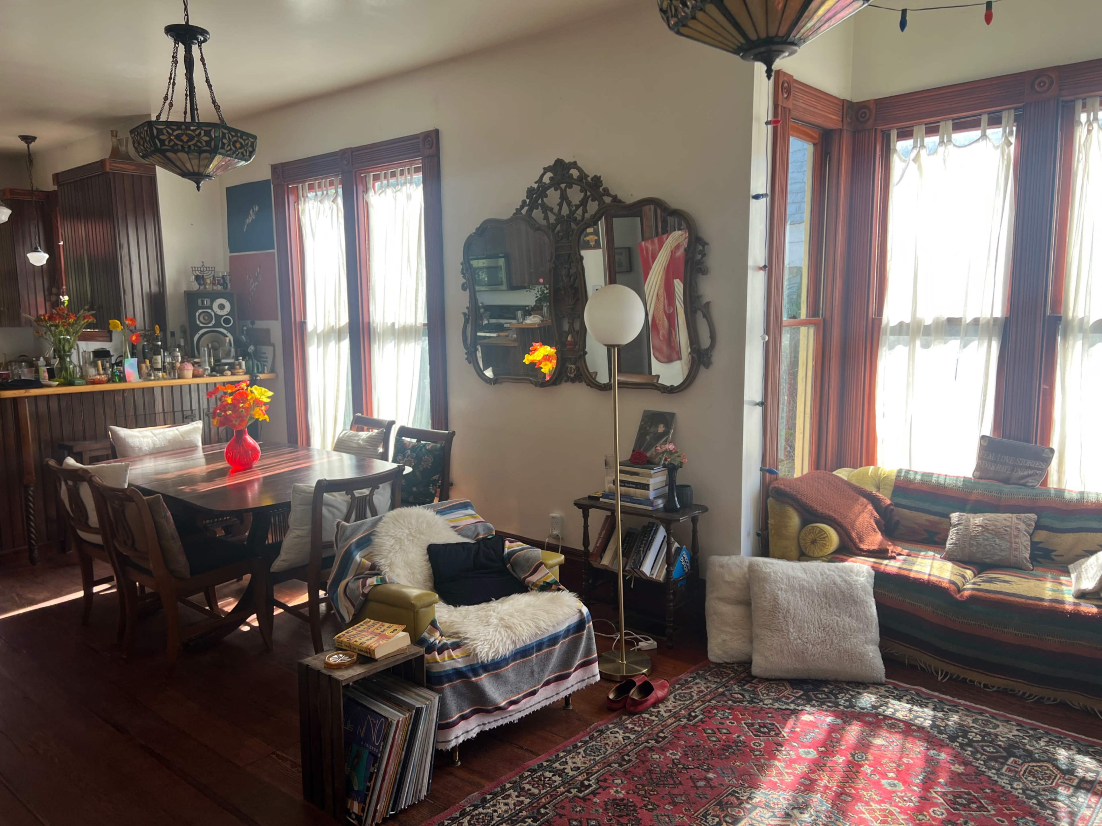 The image shows a cozy living space featuring a dining table with chairs, decorative mirrors, a vibrant rug, and a seating area with cushions and blankets.