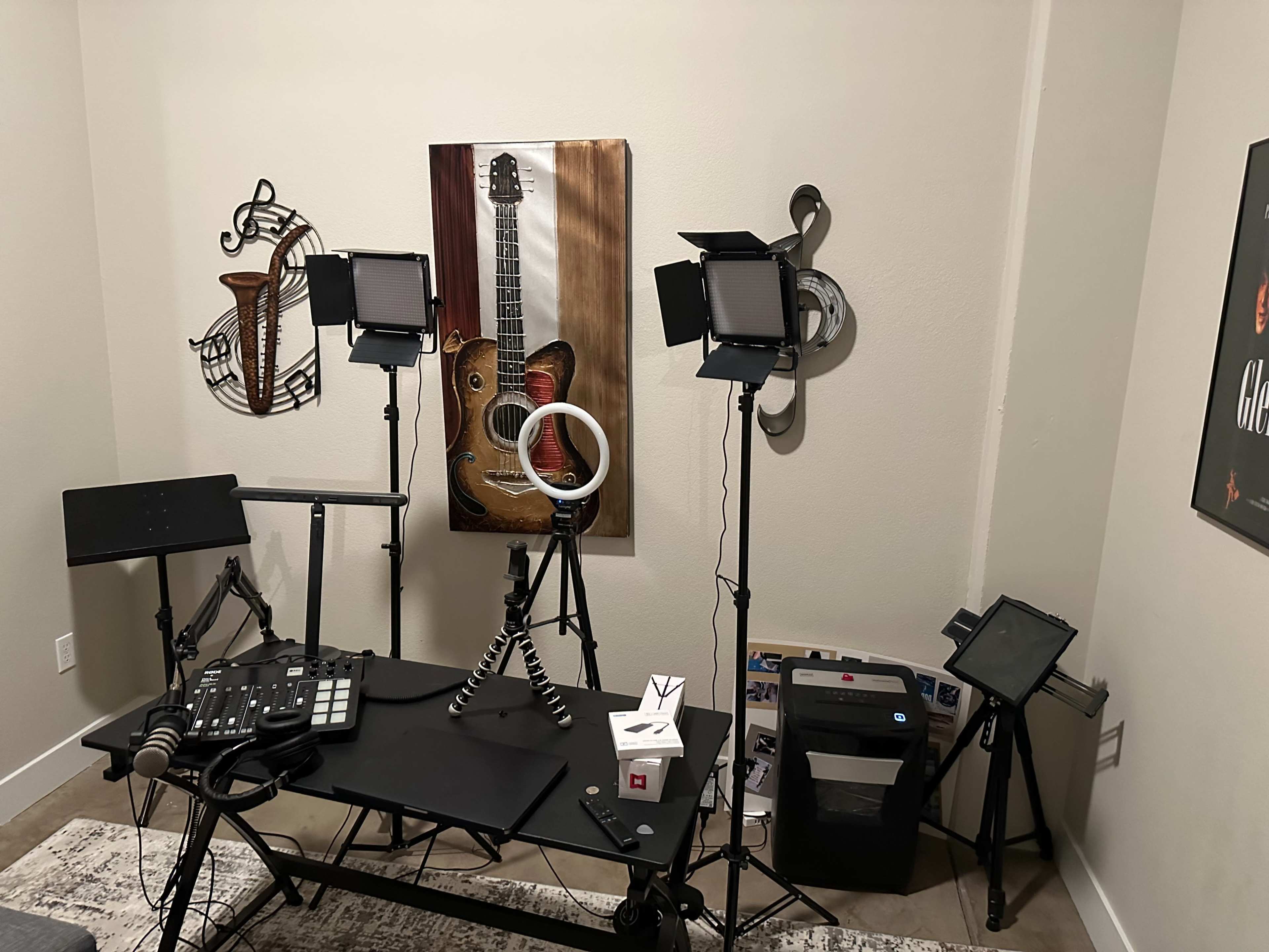 The image shows a room setup featuring a desk with a music production pad, multiple lighting sources, a tripod with a ring light, and framed musical decor on the walls.