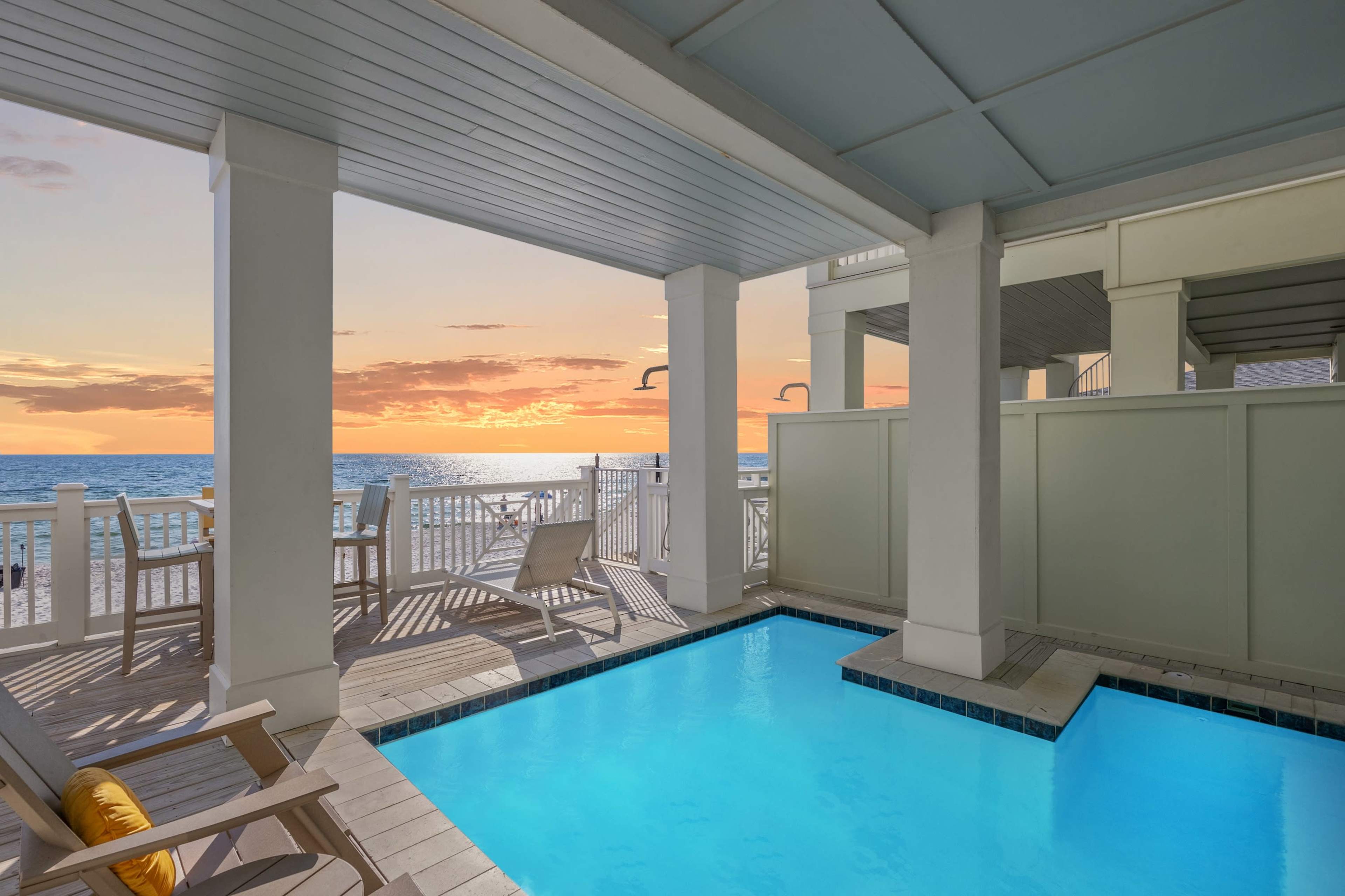 The image shows a poolside area with lounge chairs, overlooking a beach and ocean at sunset.
