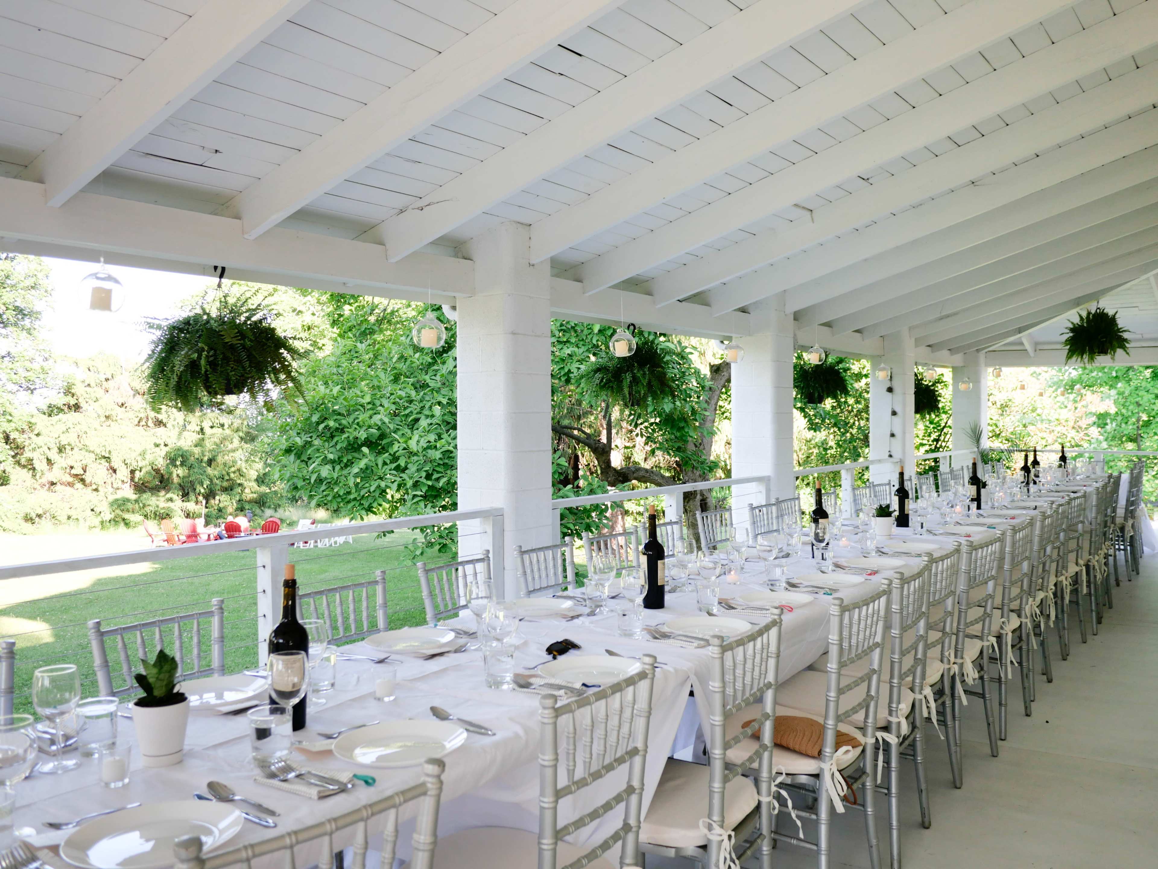 A long dining table is set up under a white, beamed patio, surrounded by greenery and decorated with potted plants and wine bottles.