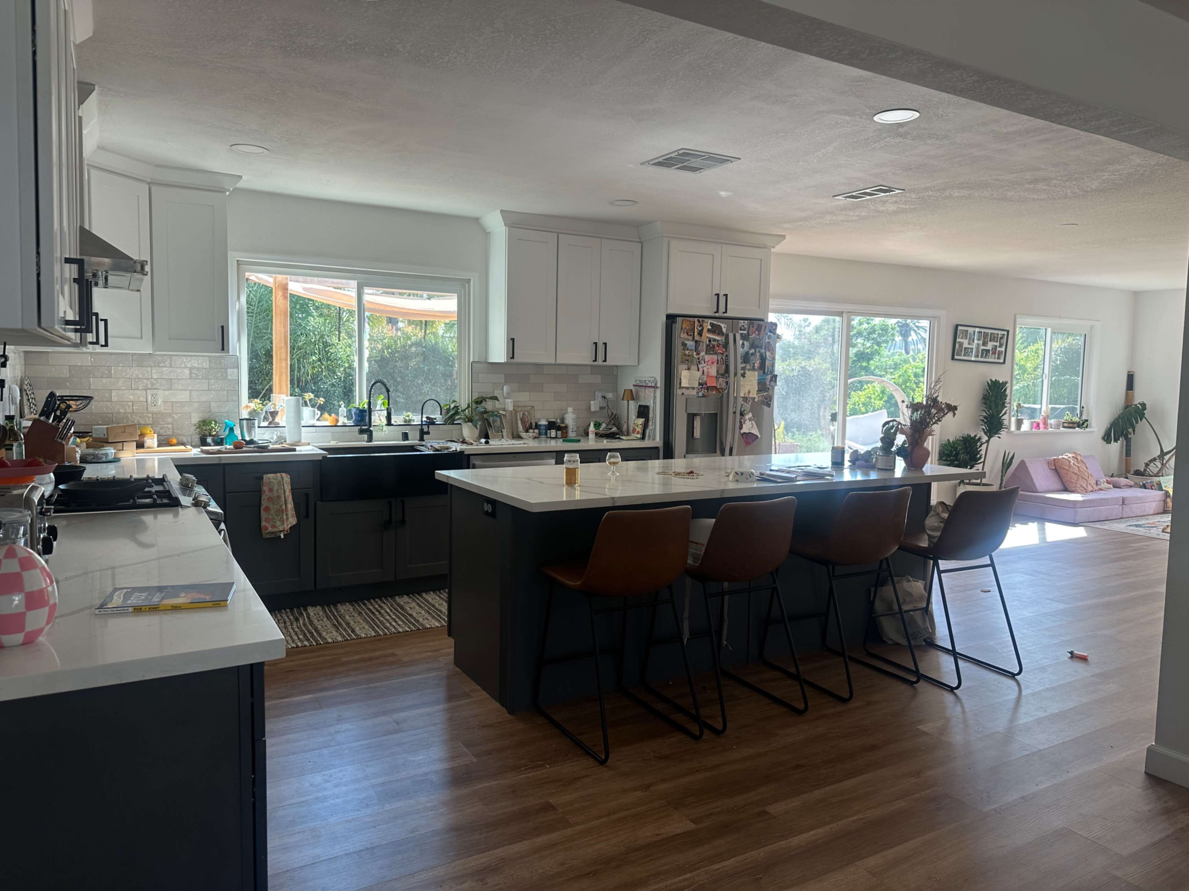 The image shows a spacious kitchen with white cabinets, a large central island with bar stools, and a view of a bright living area adorned with plants and natural light.