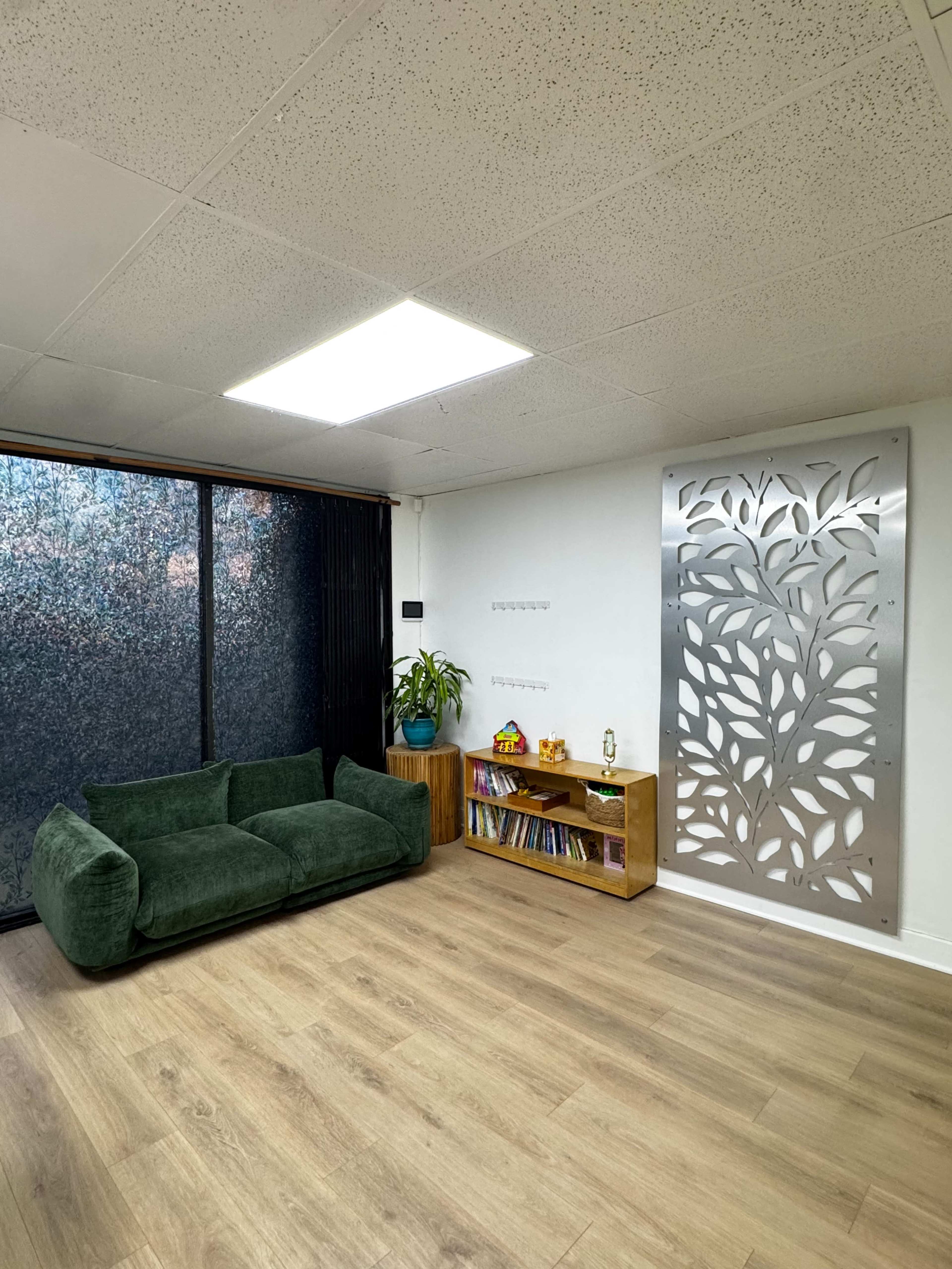 The image depicts a modern interior space featuring a green couch, a wooden shelf with books and decorative items, and a decorative metal panel on one wall.