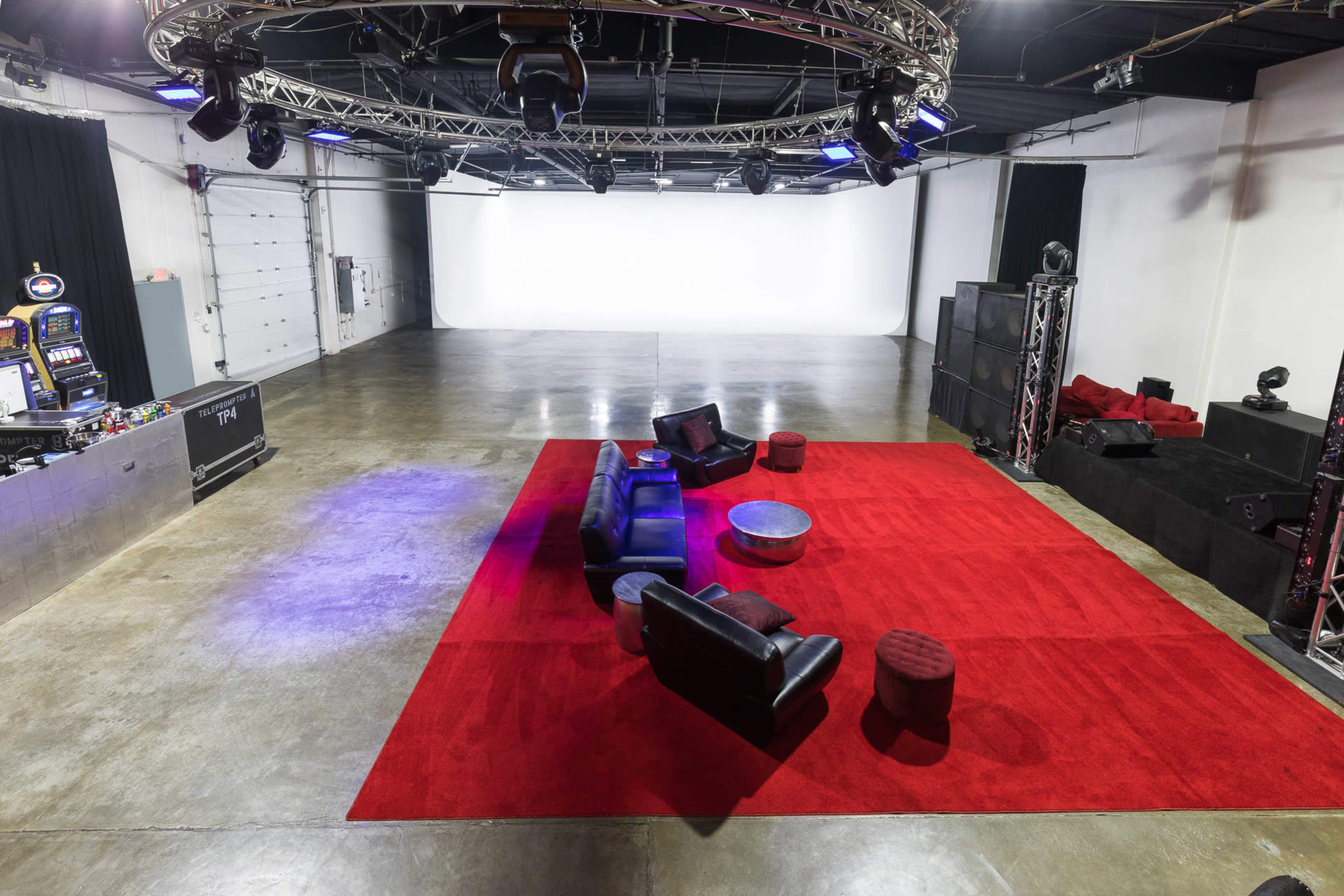The image shows an empty studio space featuring a red carpet, a black leather couch, a circular table, and a gaming machine against the wall.
