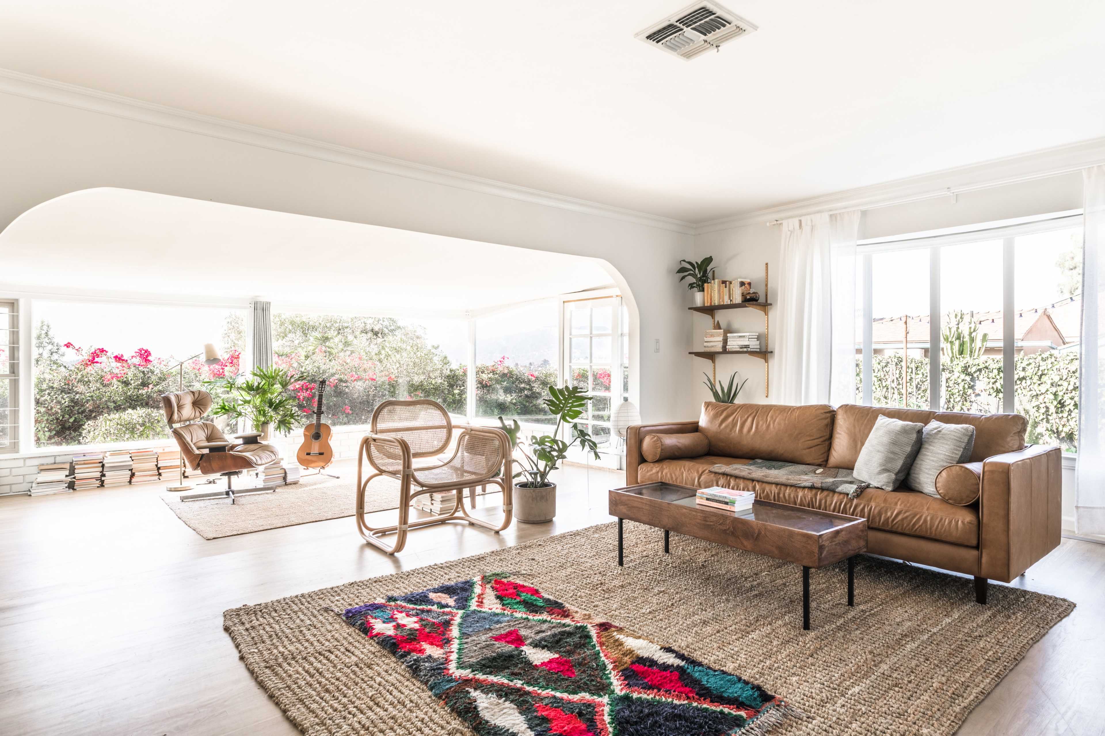 A bright, airy living room with a brown leather sofa, a wooden coffee table, a patterned rug, and a view of a well-lit space featuring plants and an acoustic guitar.