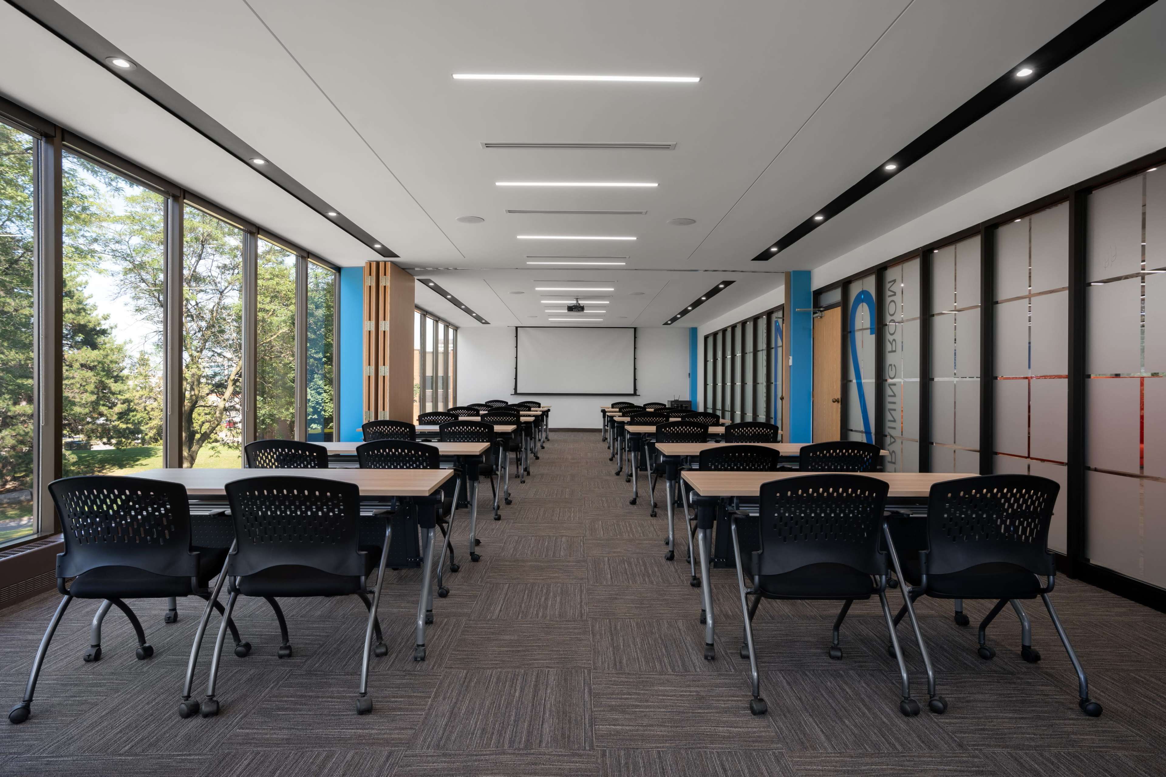 A modern classroom features rows of black chairs and tables, large windows allowing natural light, and a projector screen at the front.