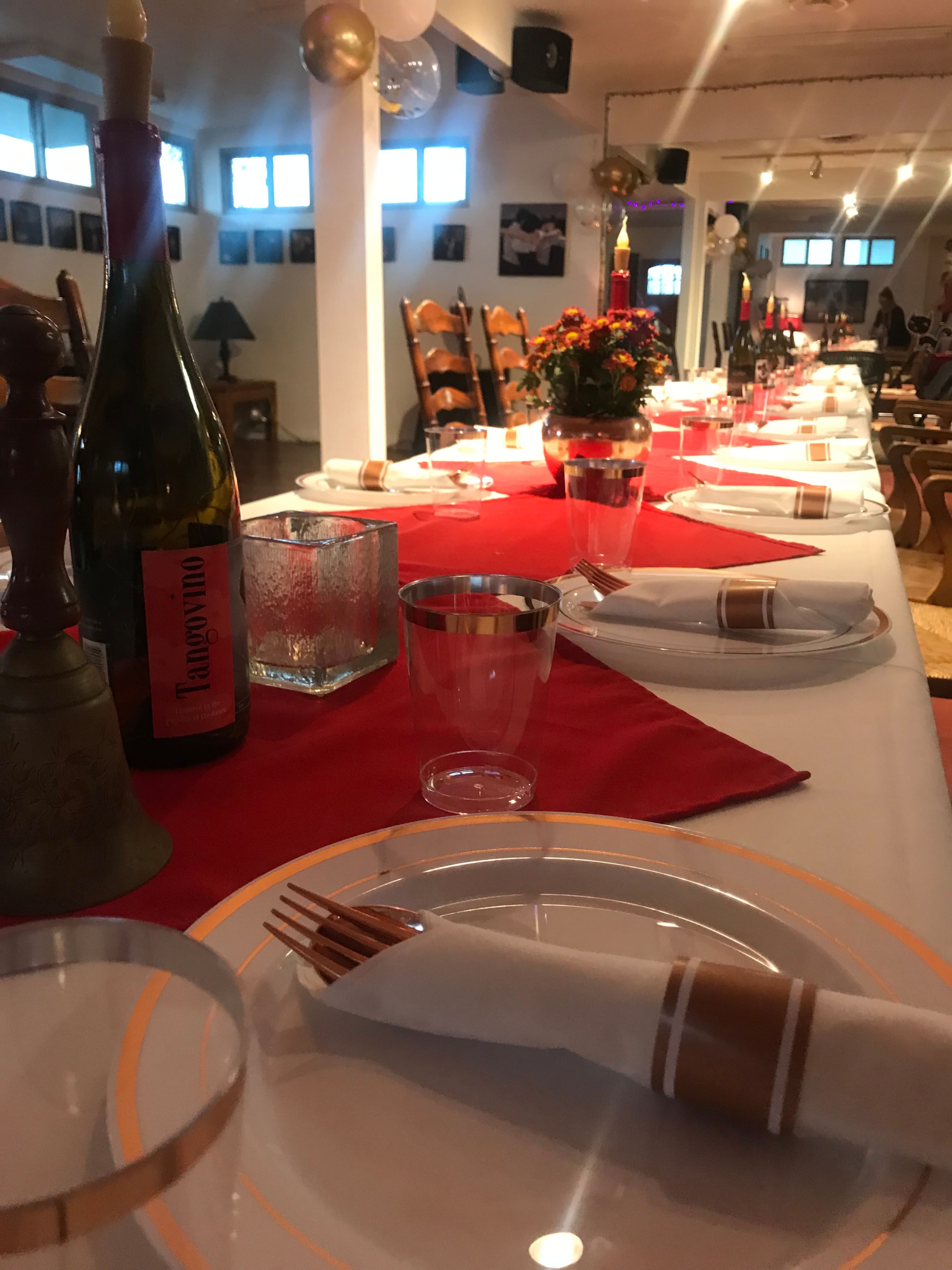 A long dining table is set with plates, utensils, and a bottle of wine, decorated with a red tablecloth and flowers in the background.