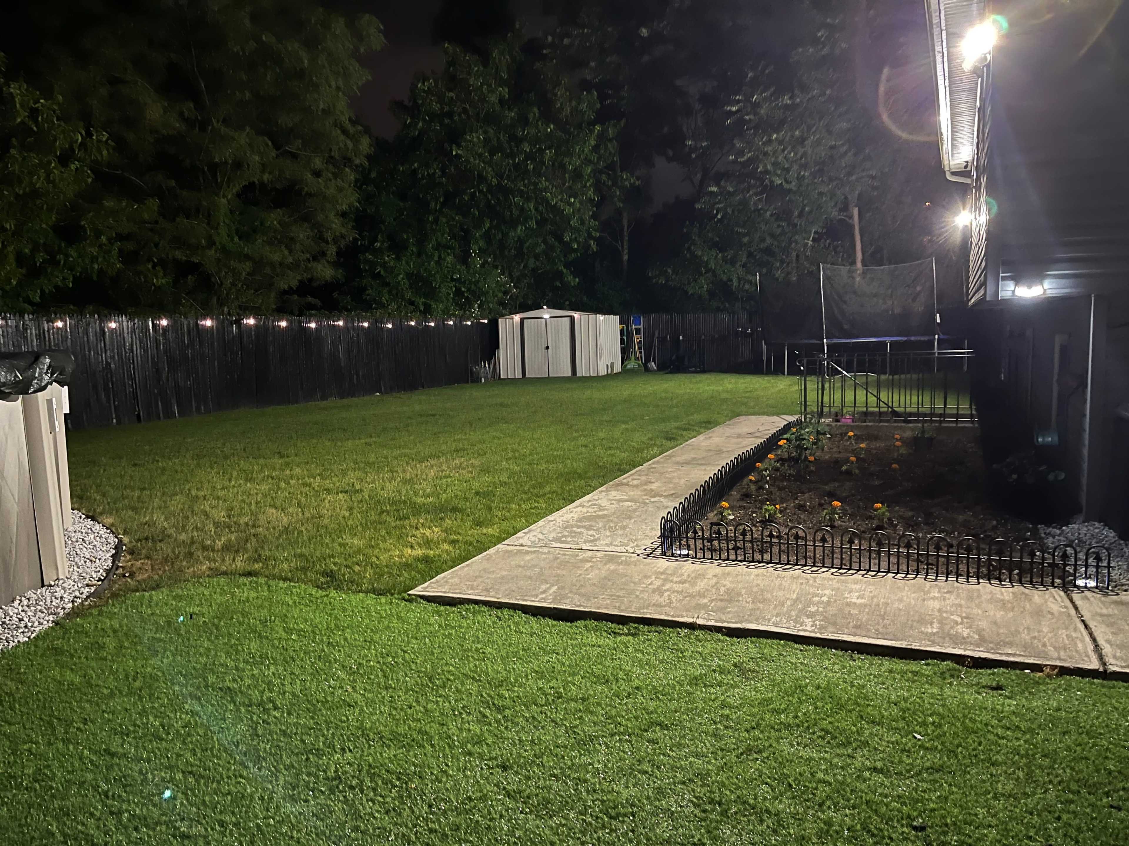 A well-maintained backyard features a neatly mowed lawn, a garden with flower beds, a concrete walkway, and a shed in the background under night lighting.