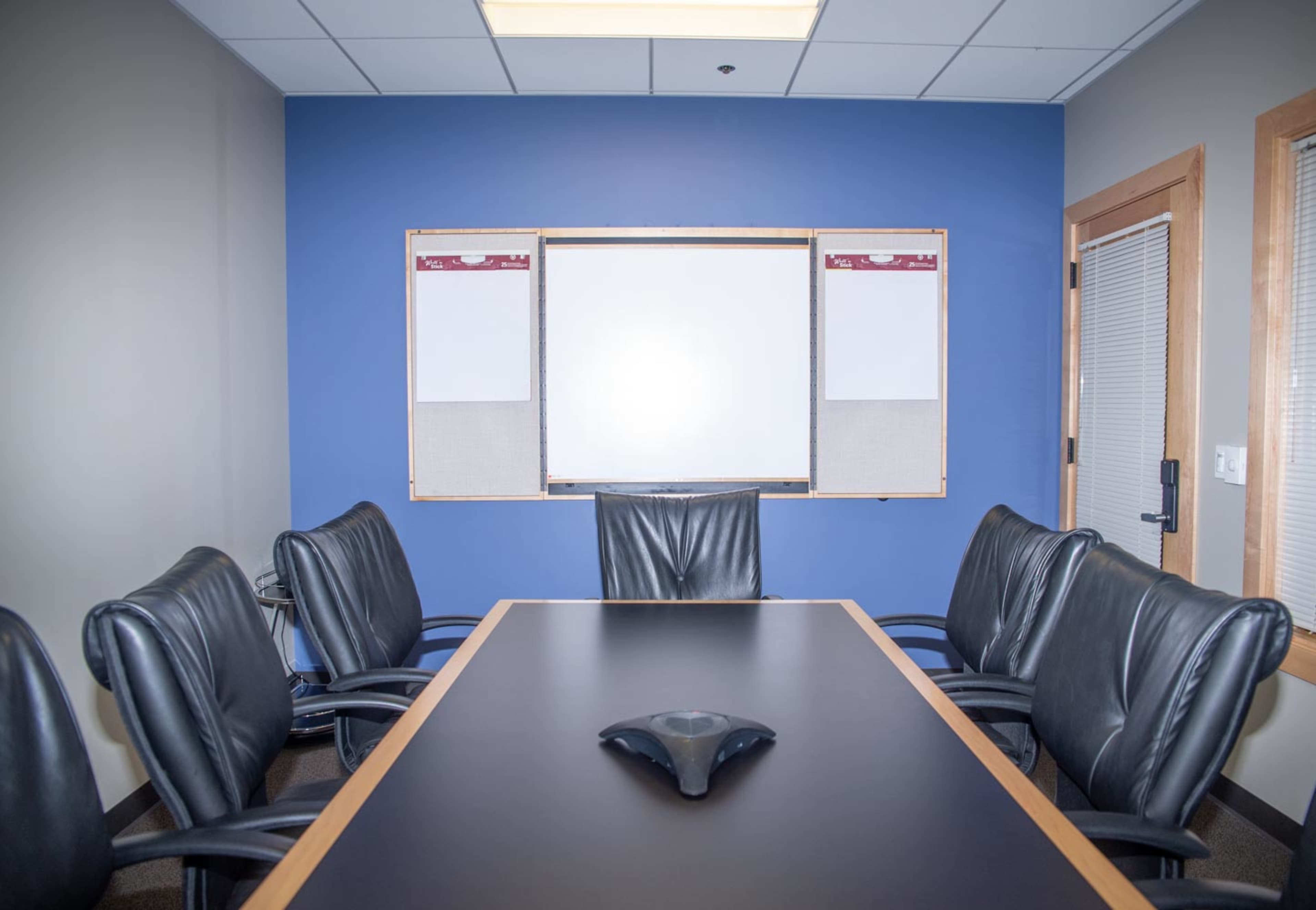 A conference room features a large table surrounded by black leather chairs, with a whiteboard and two bulletin boards mounted on a blue wall.