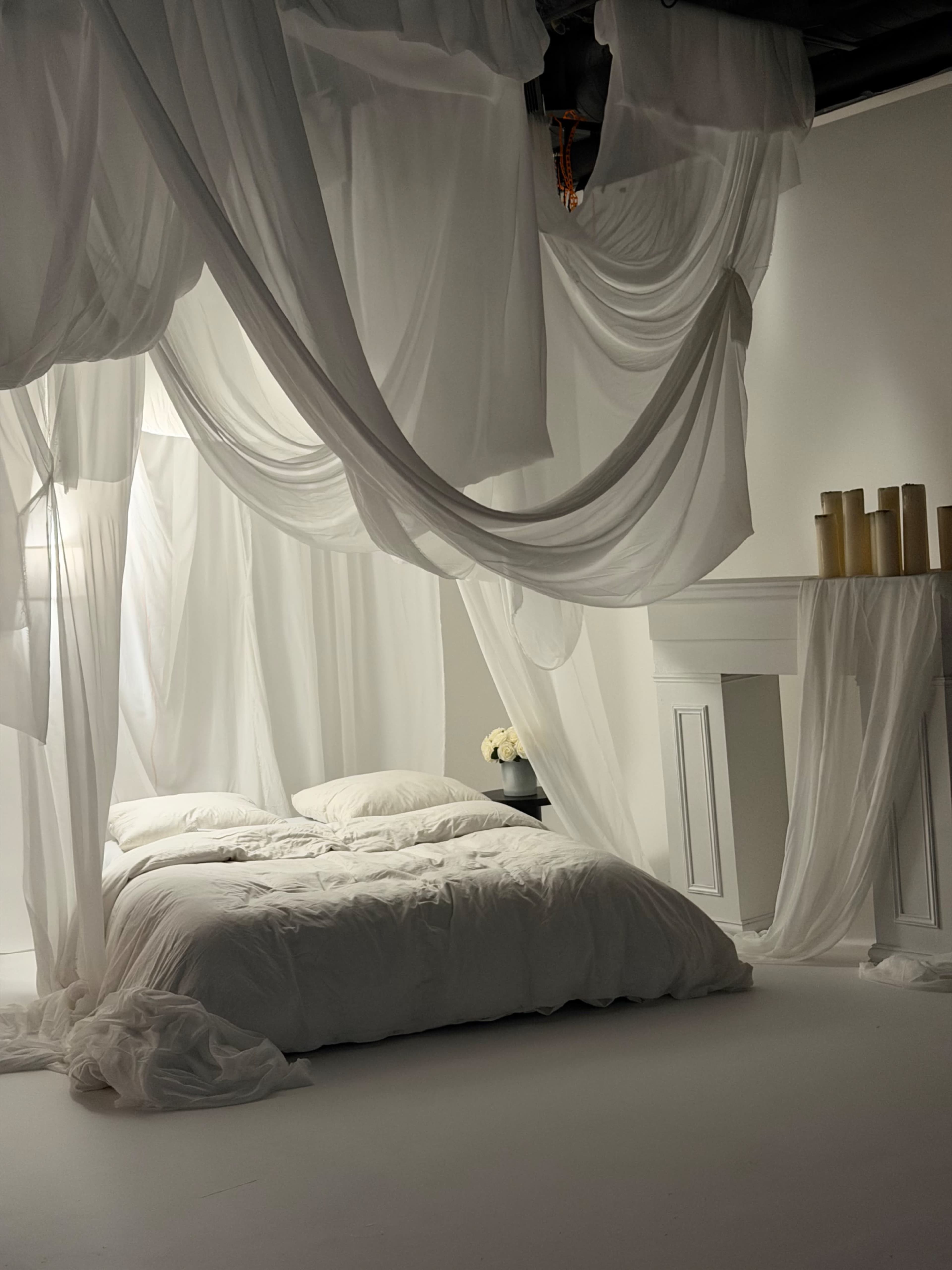 A neatly arranged bed with white linens is surrounded by flowing white drapery in a softly lit room.
