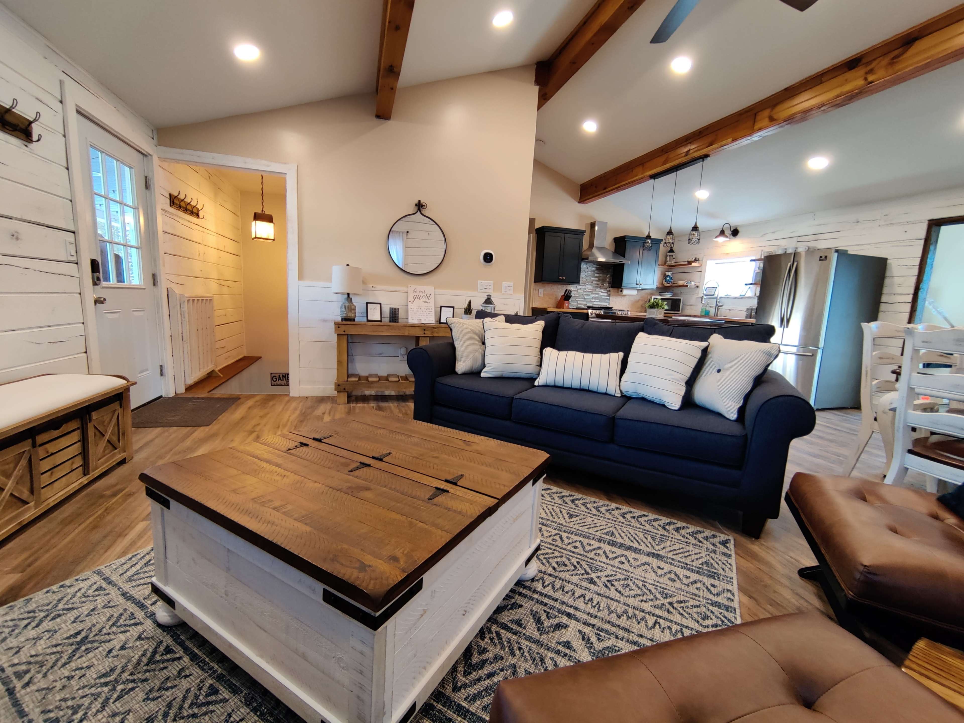 The image shows a cozy living room with a blue couch, a wooden coffee table, and a dining area, featuring light wooden beams and white walls.