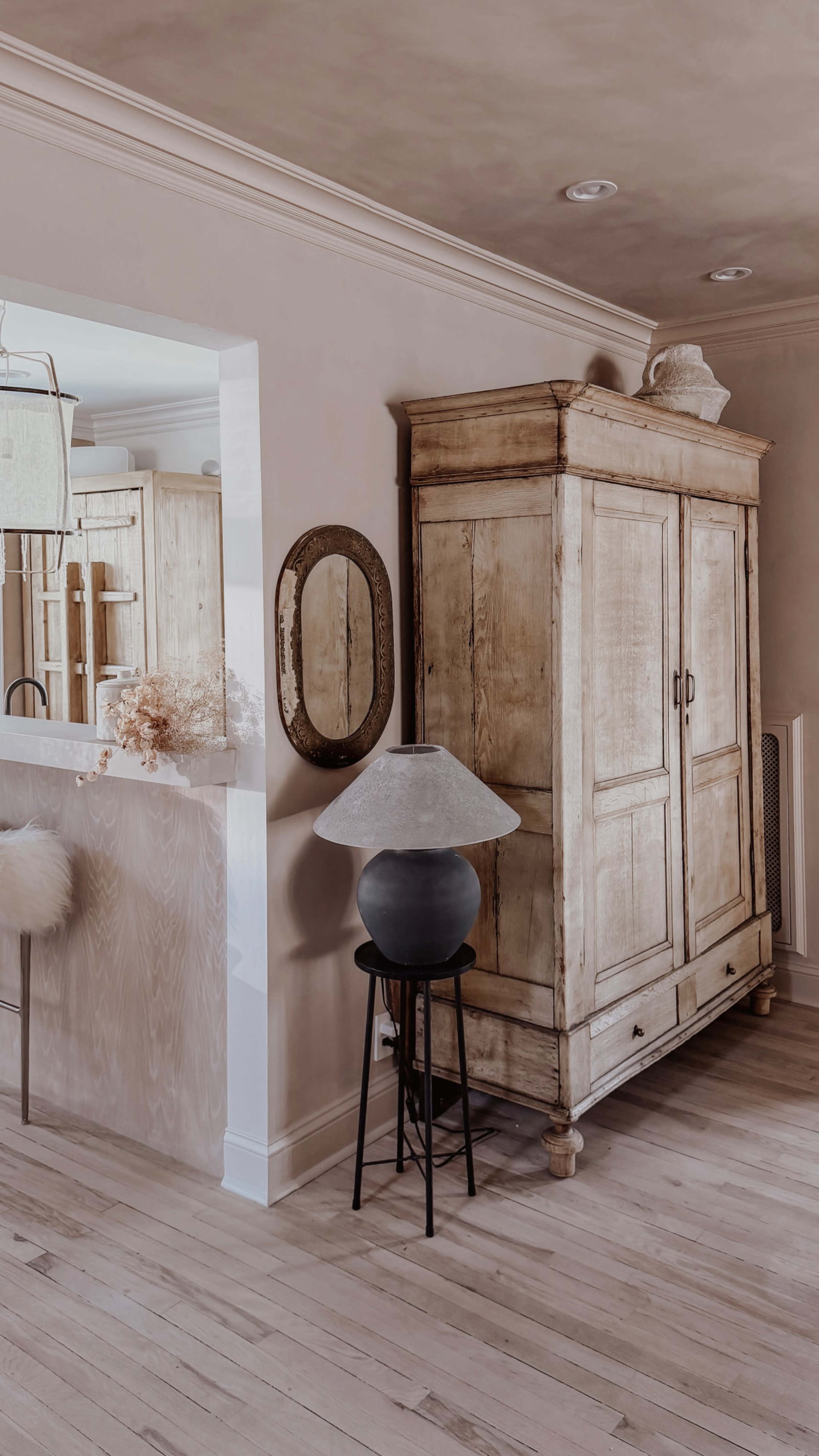 The image shows a light-colored interior featuring a wooden armoire, a round mirror, and a lamp on a black stool, with a glimpse of a kitchen in the background.