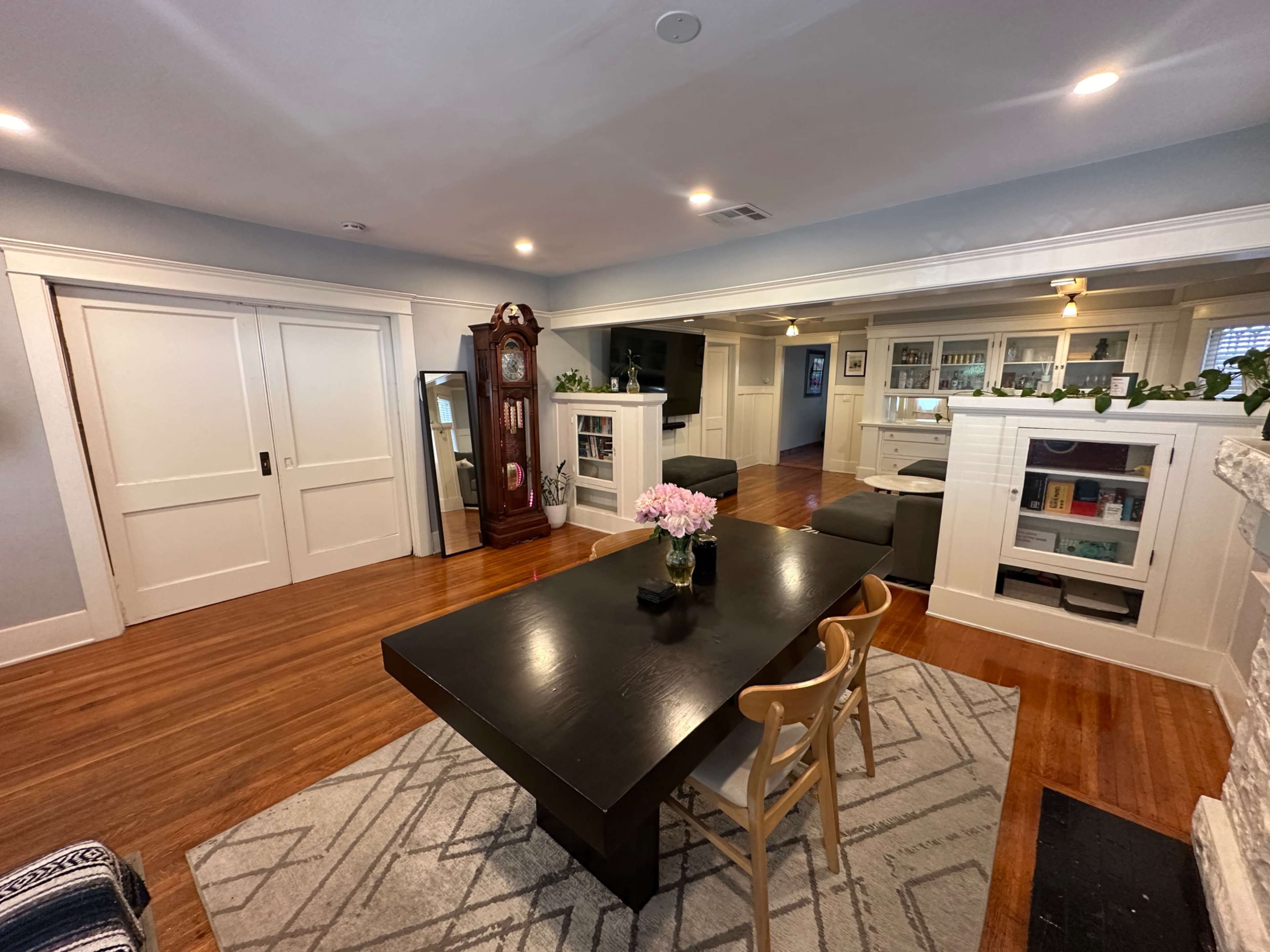 The image shows a well-lit room featuring a dining table with chairs, a decorative clock, a television, and built-in shelves against the walls.