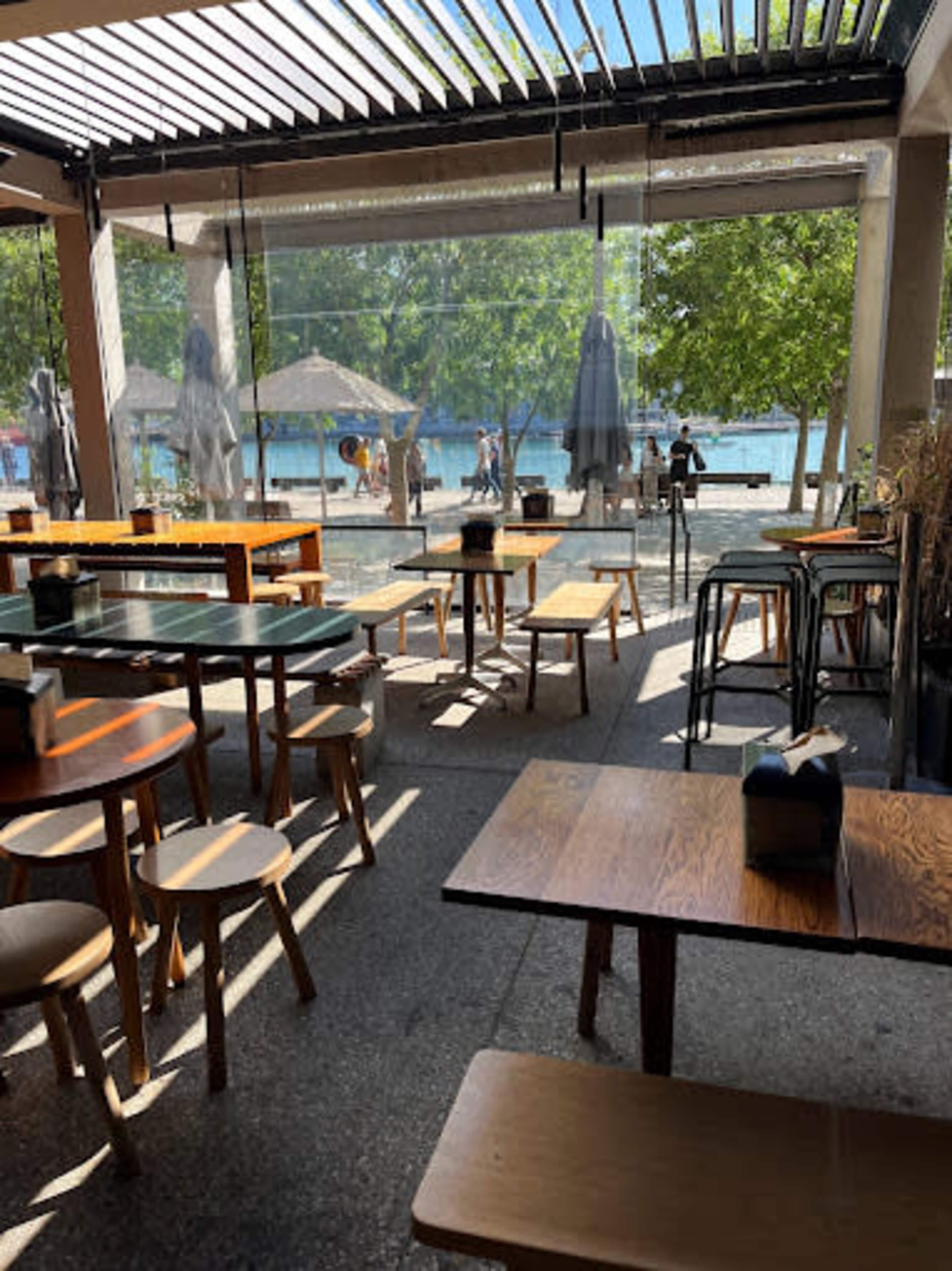 The image shows an outdoor dining area of a restaurant with wooden tables and chairs, surrounded by greenery and a view of a body of water.