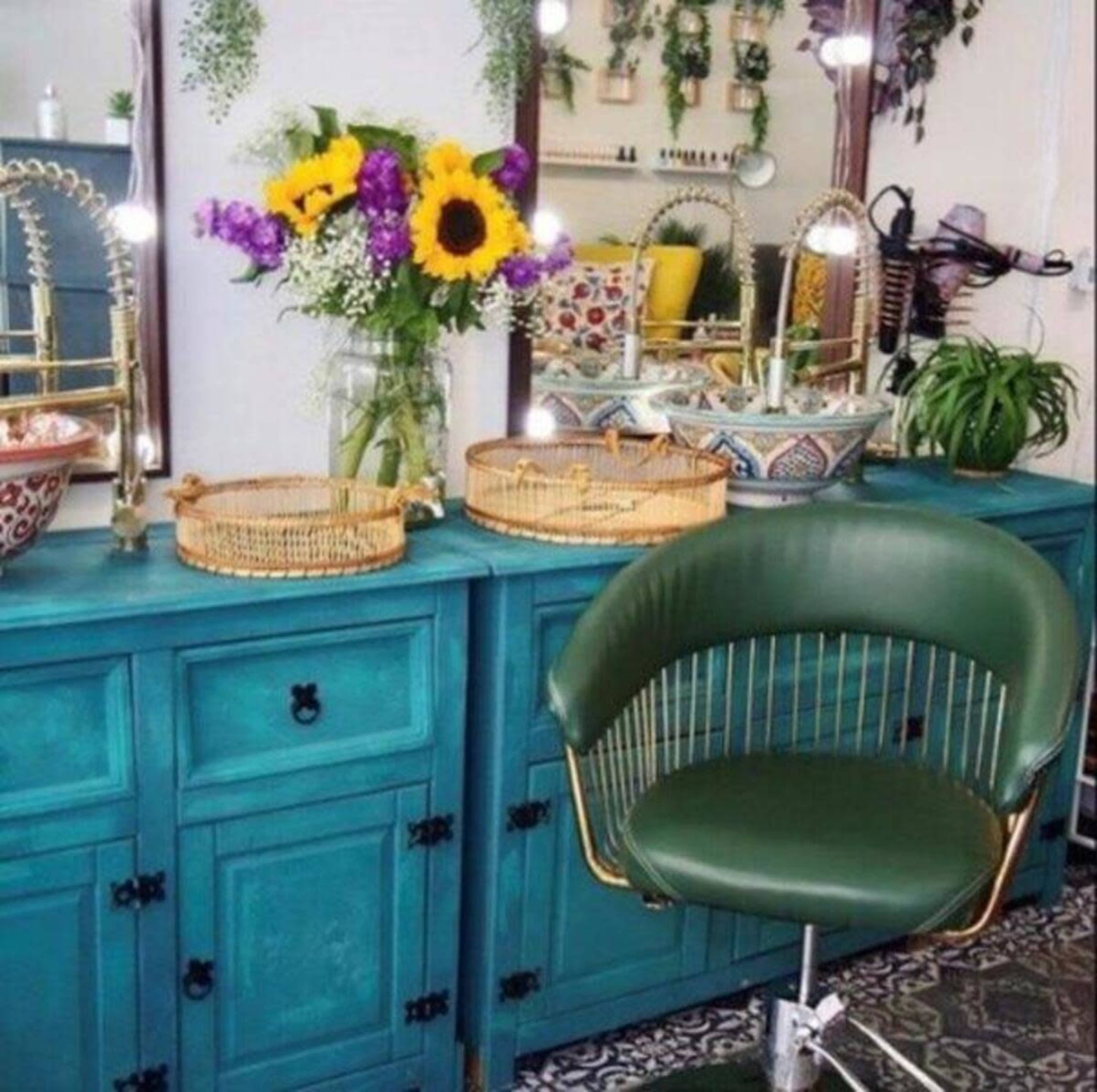 A turquoise cabinet with a green chair and a vase of sunflowers on a countertop, surrounded by mirrors and decorative items.