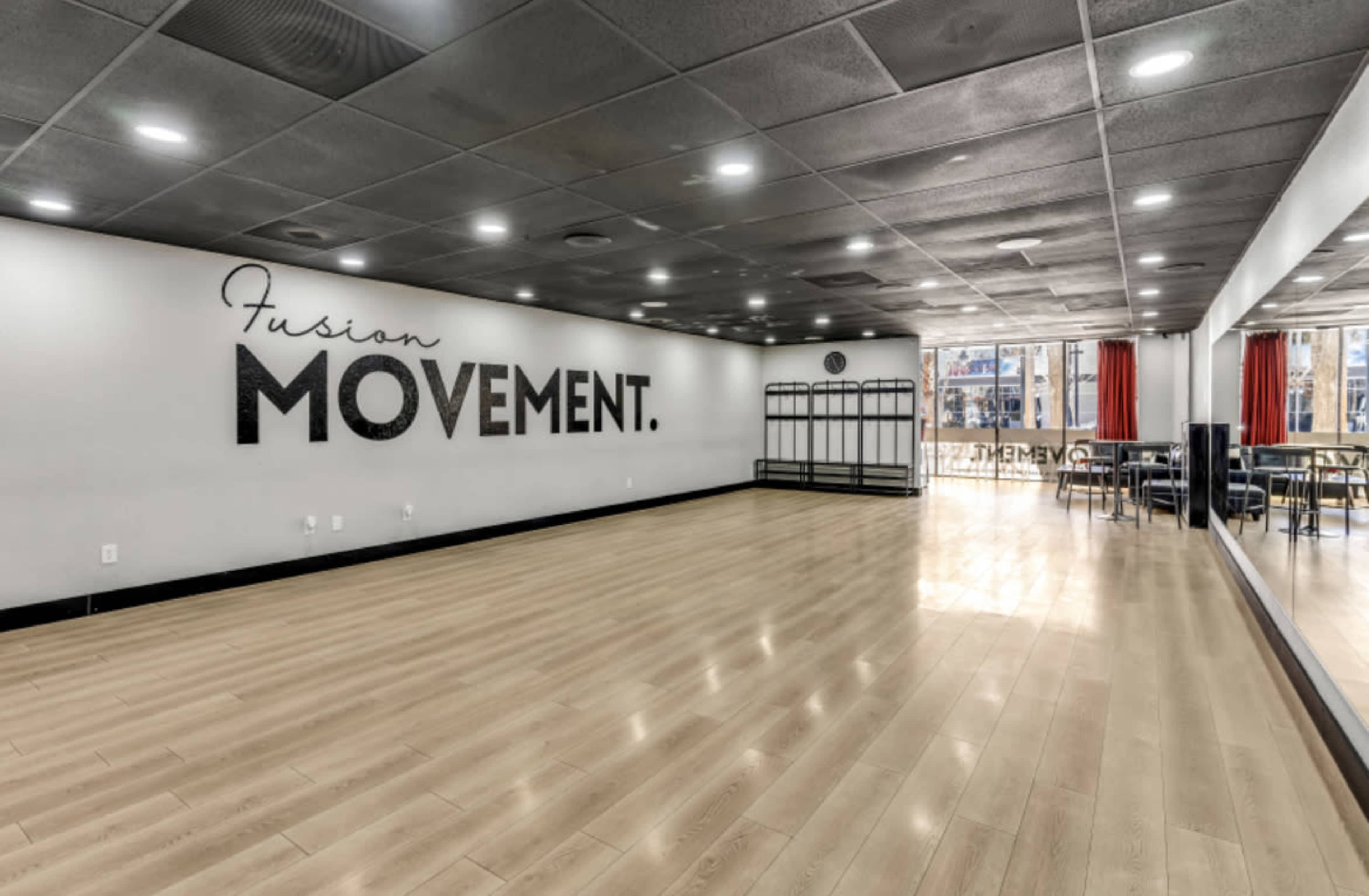 The image shows a spacious dance studio with wooden flooring, large windows, and the words "Fusion MOVEMENT." displayed on the wall.