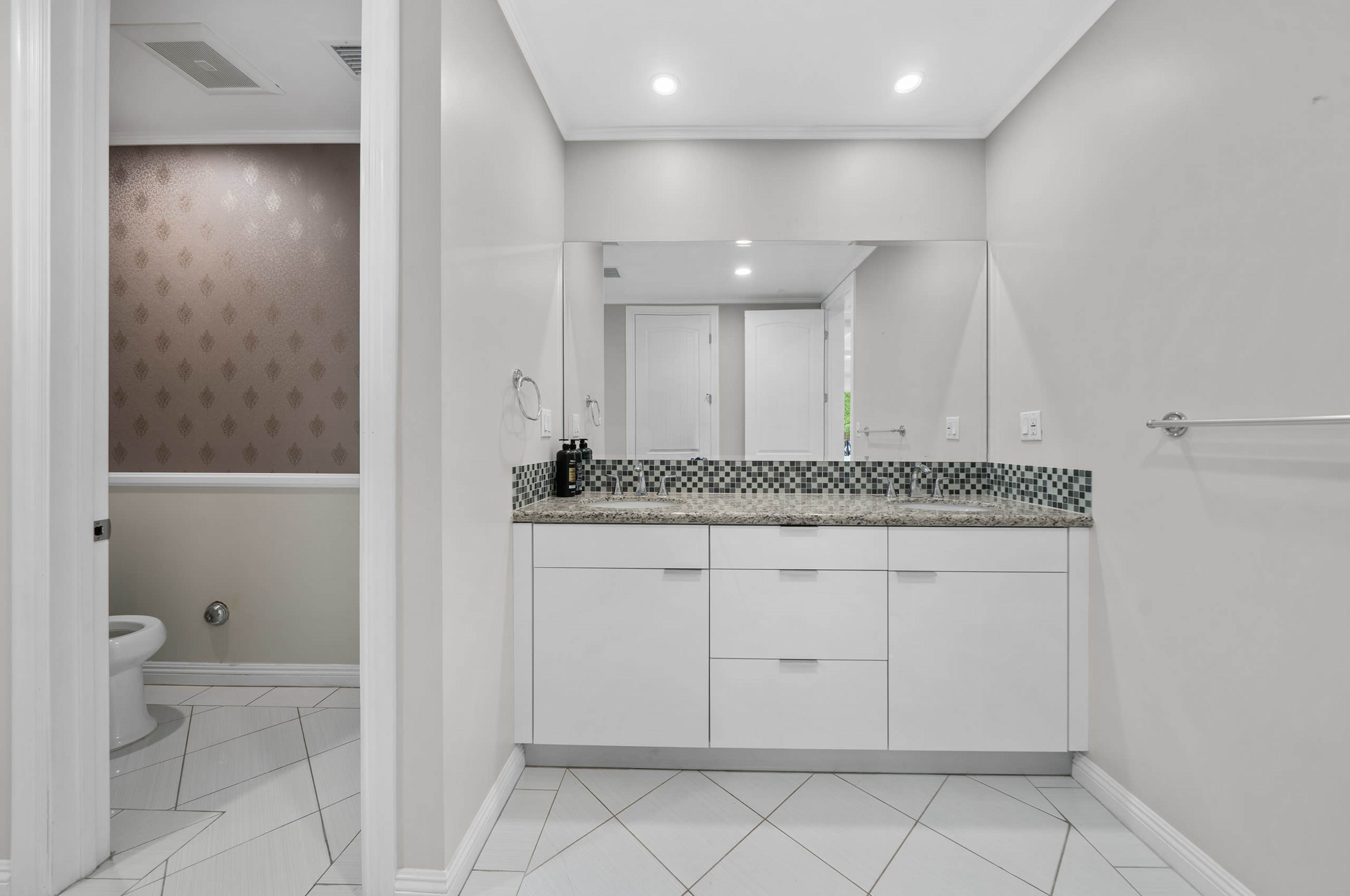 The image shows a modern bathroom with a double vanity, large mirror, and a toilet in a separate alcove.