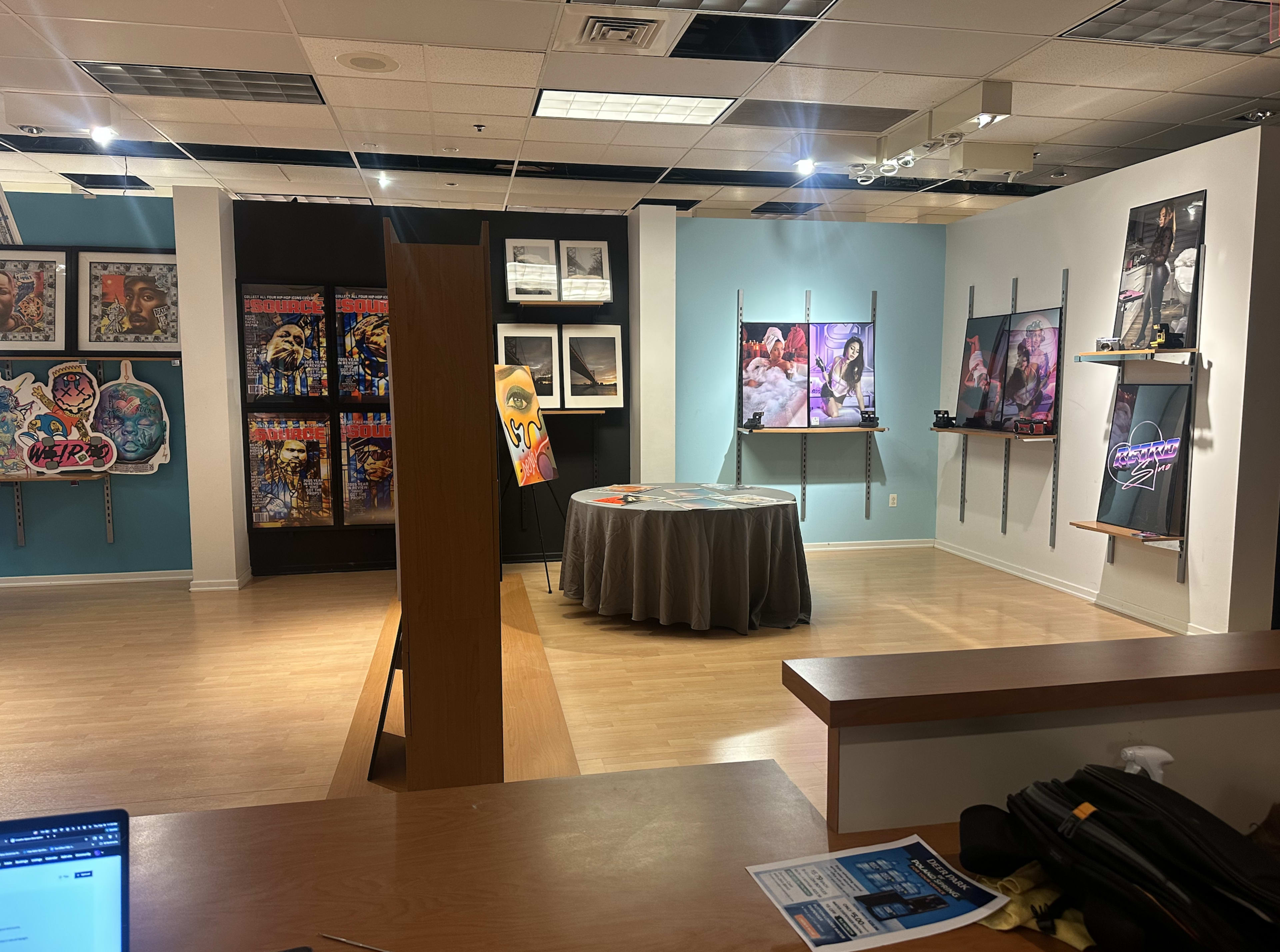 The image shows an art gallery featuring various framed artworks on the walls, a central circular table covered with a grey tablecloth, and display panels with colorful graphics.
