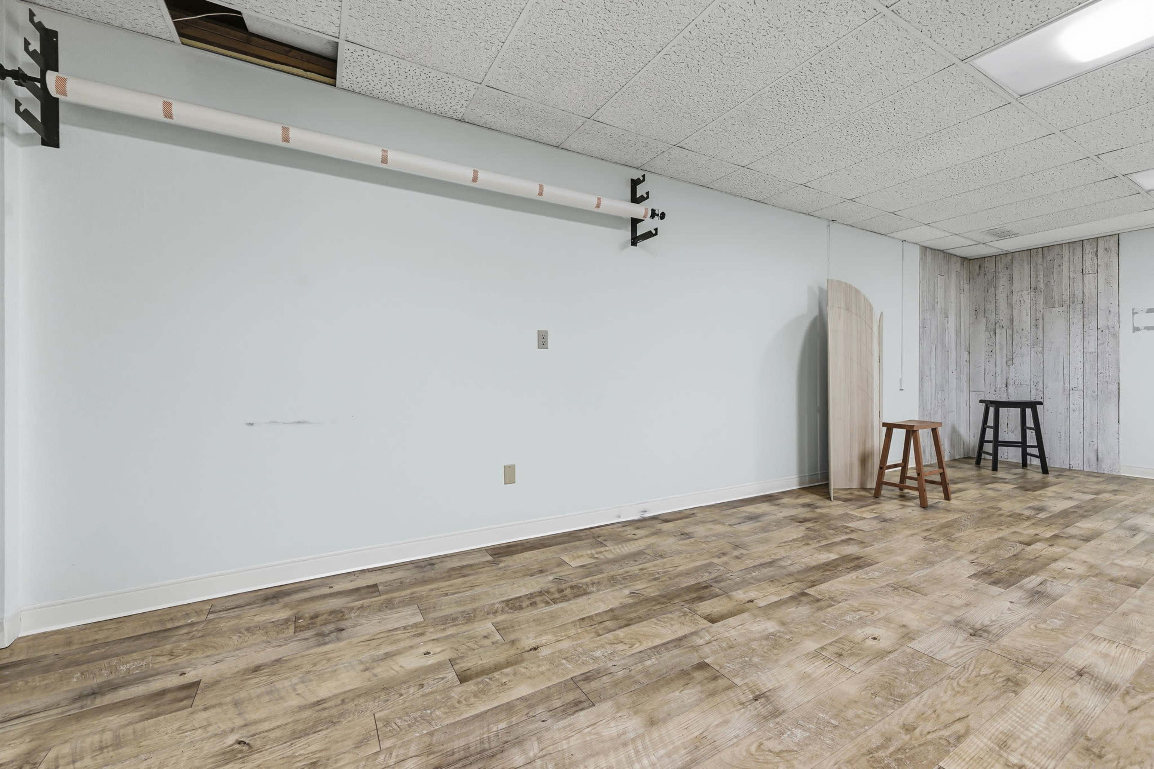The image shows an empty room with wooden flooring, a mounted horizontal bar along one wall, and two stools positioned against the opposite wall.