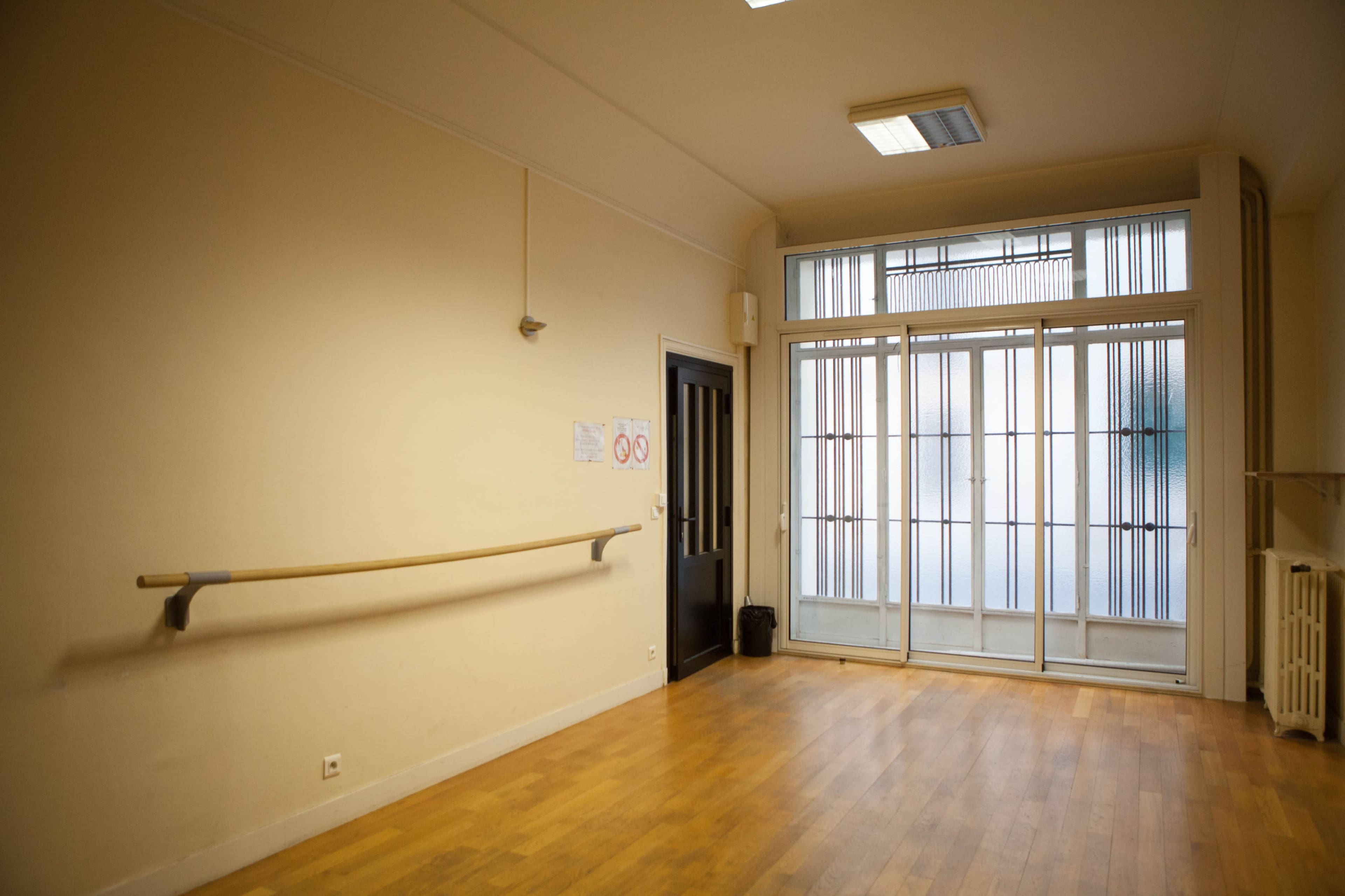 The image shows an empty dance studio with wooden flooring and large windows allowing natural light.