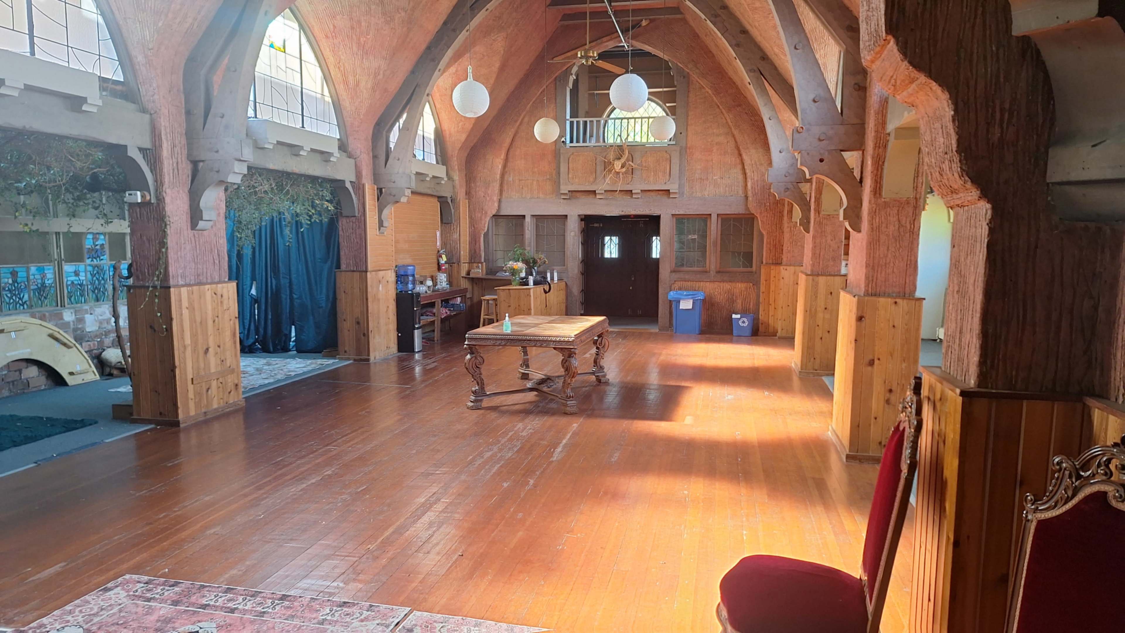 The image shows a spacious, empty interior of a building with wooden beams, a polished wooden floor, and a large table at the center.
