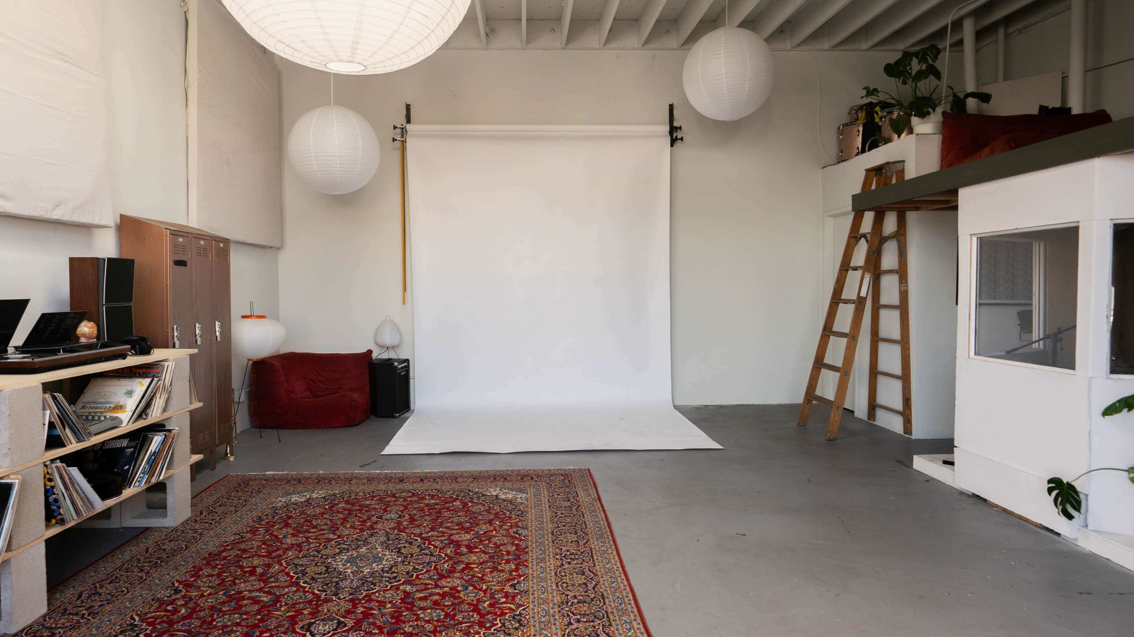 A spacious studio with a white backdrop, a wooden ladder, a patterned rug, and several hanging paper lanterns.
