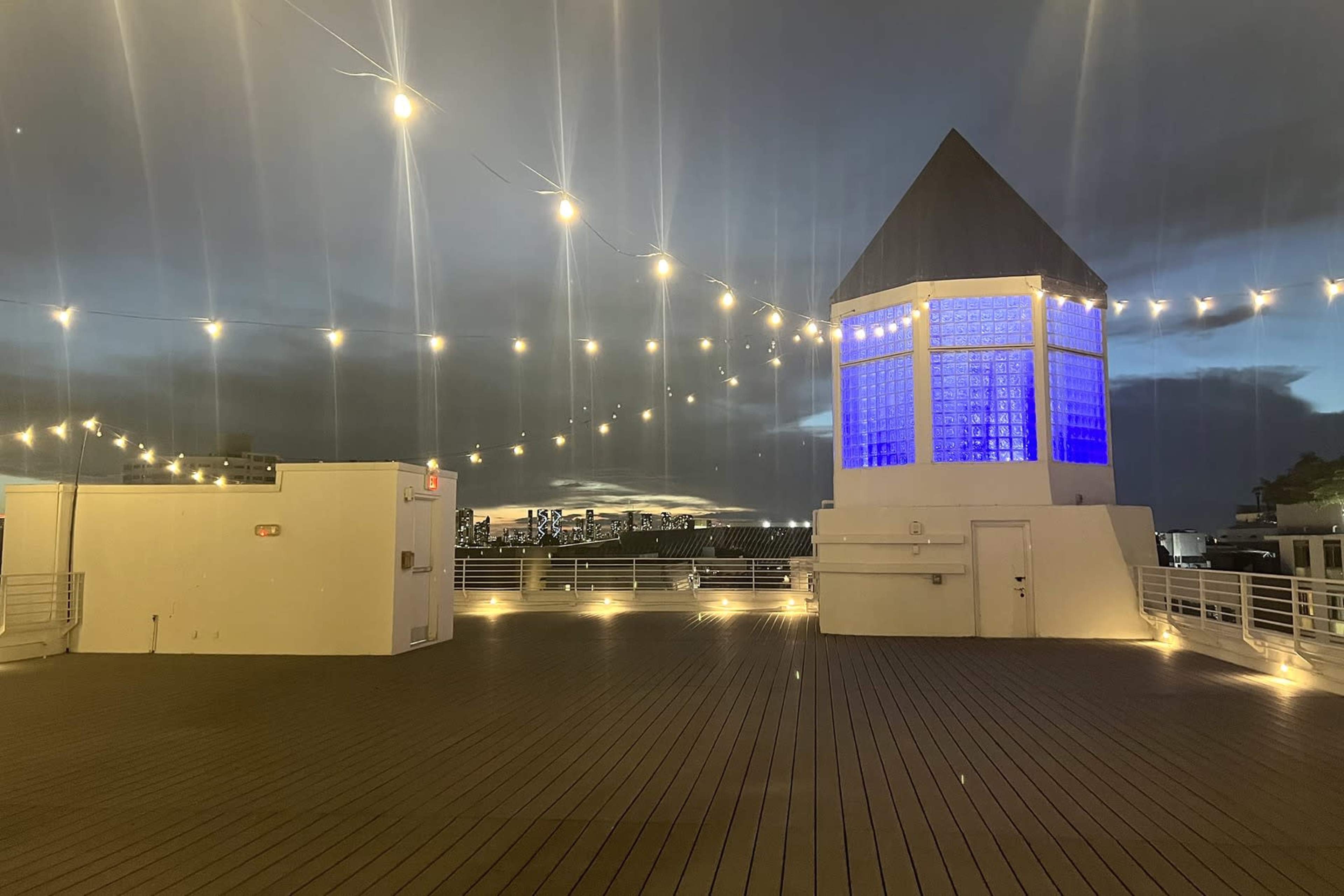 The image shows a rooftop area featuring a small tower with blue lighting and string lights illuminating the space at dusk.