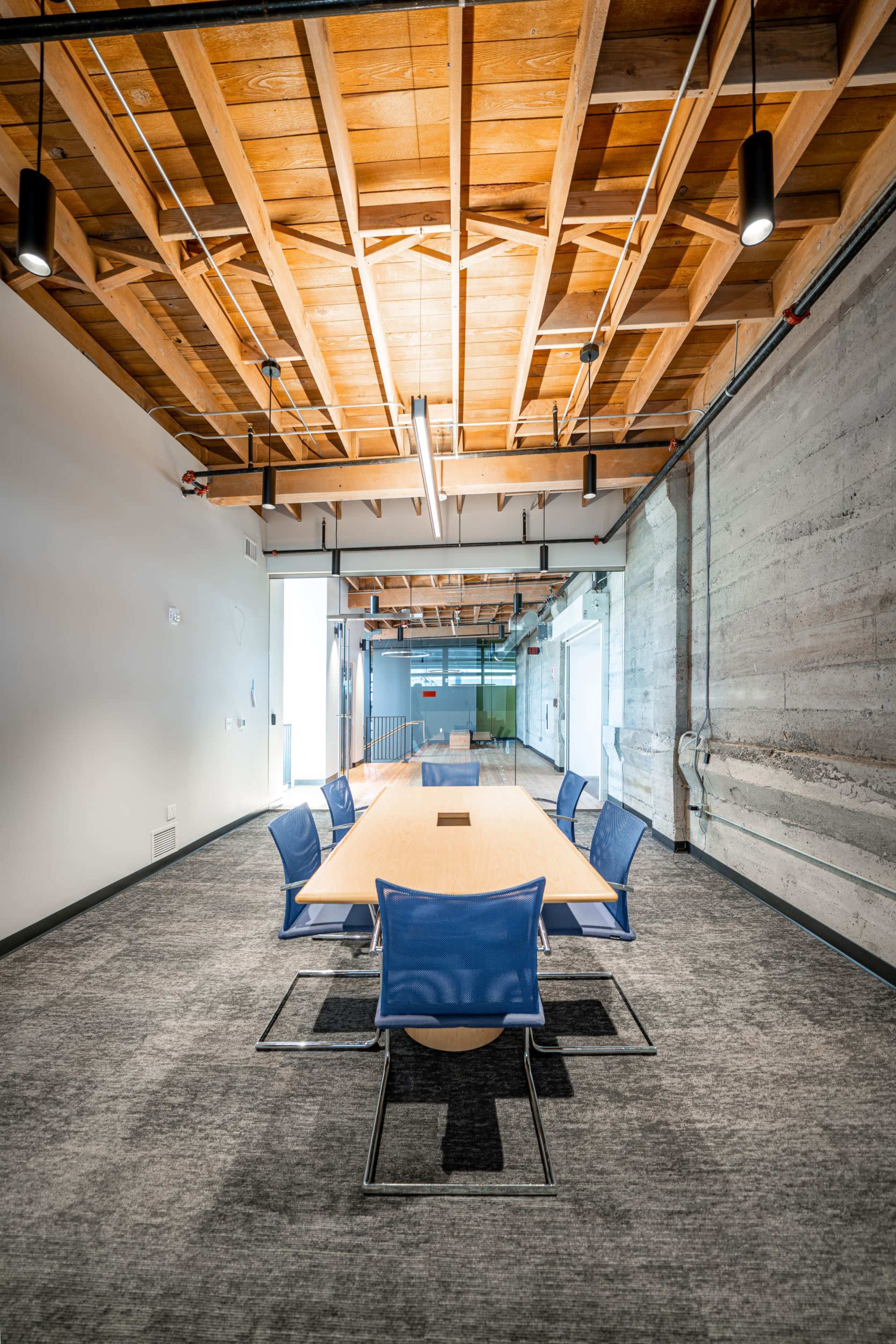 A conference room features a long wooden table surrounded by blue chairs, with exposed wooden beams and concrete walls.