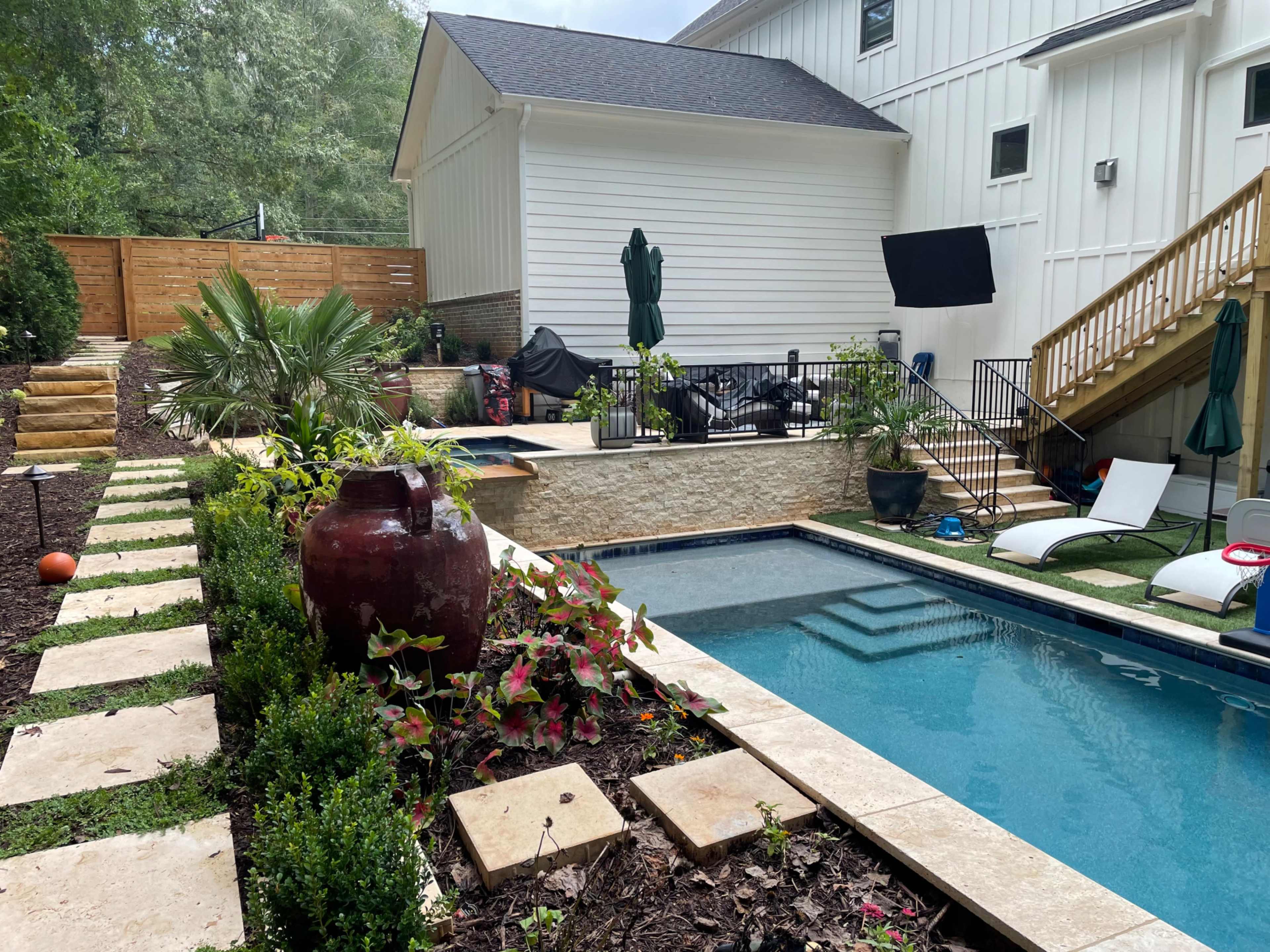 The image shows a backyard with a swimming pool surrounded by landscaping, stone pathways, and a nearby house.