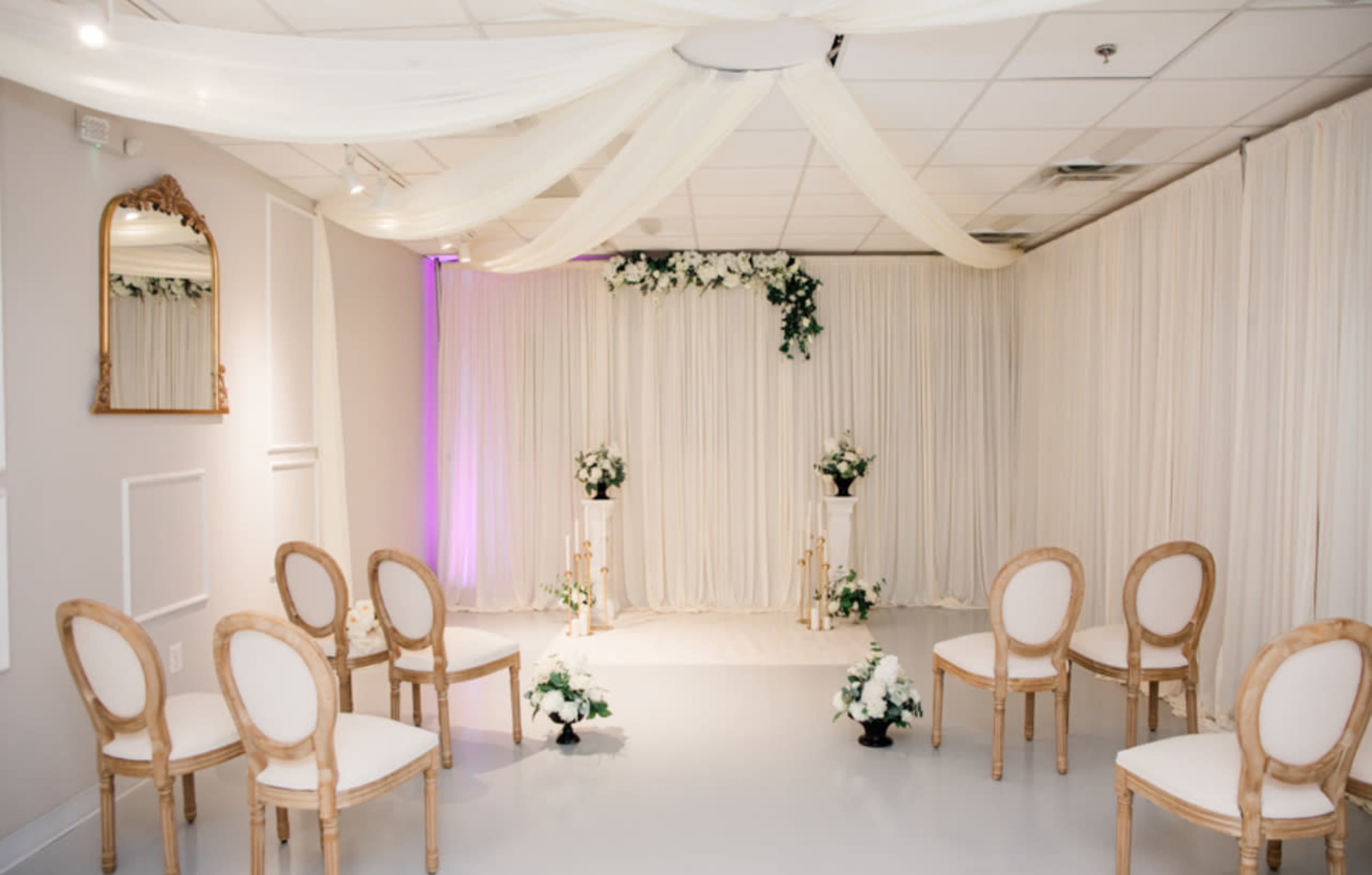 The image shows a softly lit event space with white drapes, floral arrangements, and wooden chairs arranged in a semicircle facing an altar area adorned with more flowers.