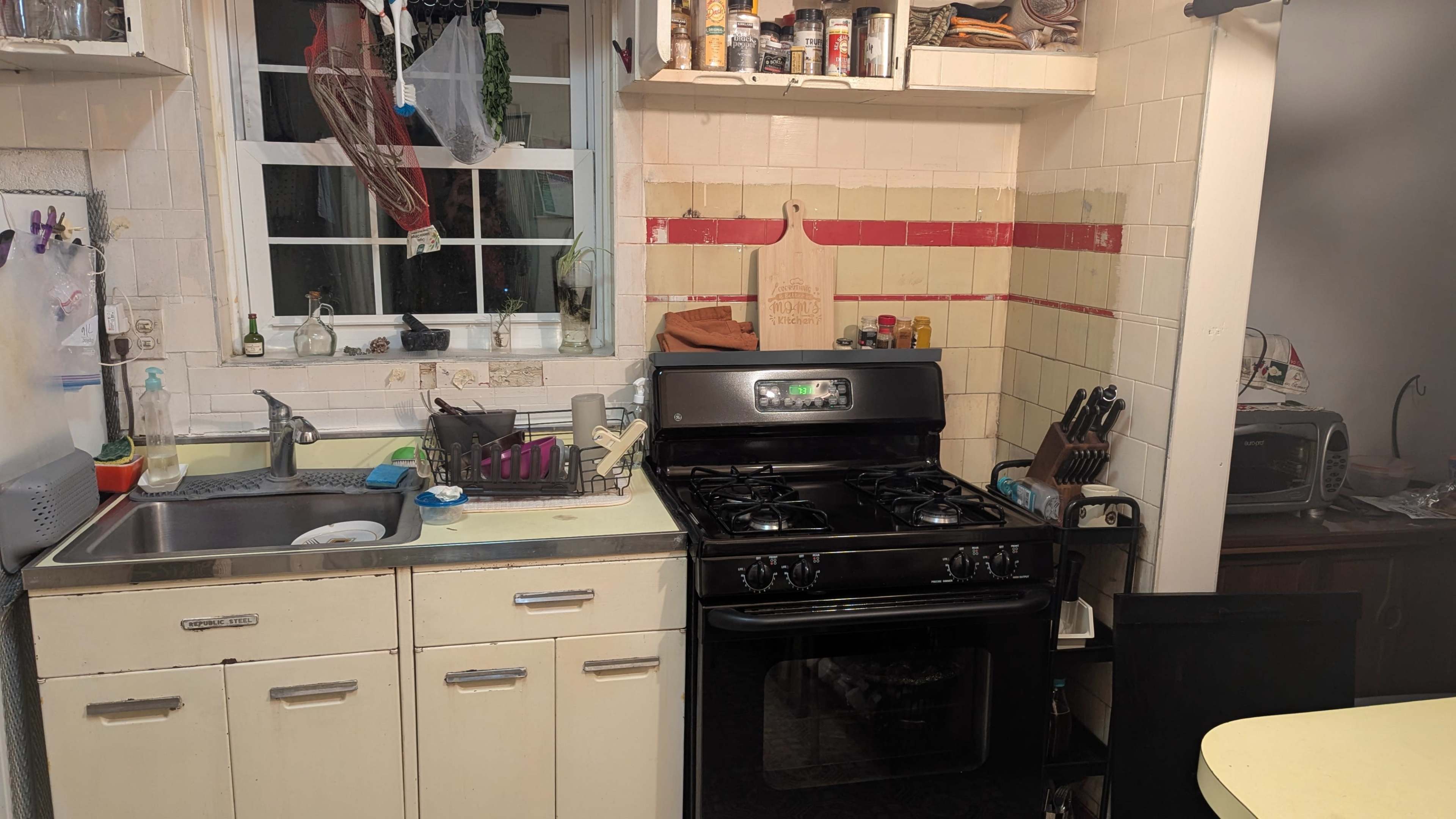 The image shows a small kitchen with a black gas stove, a stainless steel sink, and various kitchen items on the countertop and shelves.