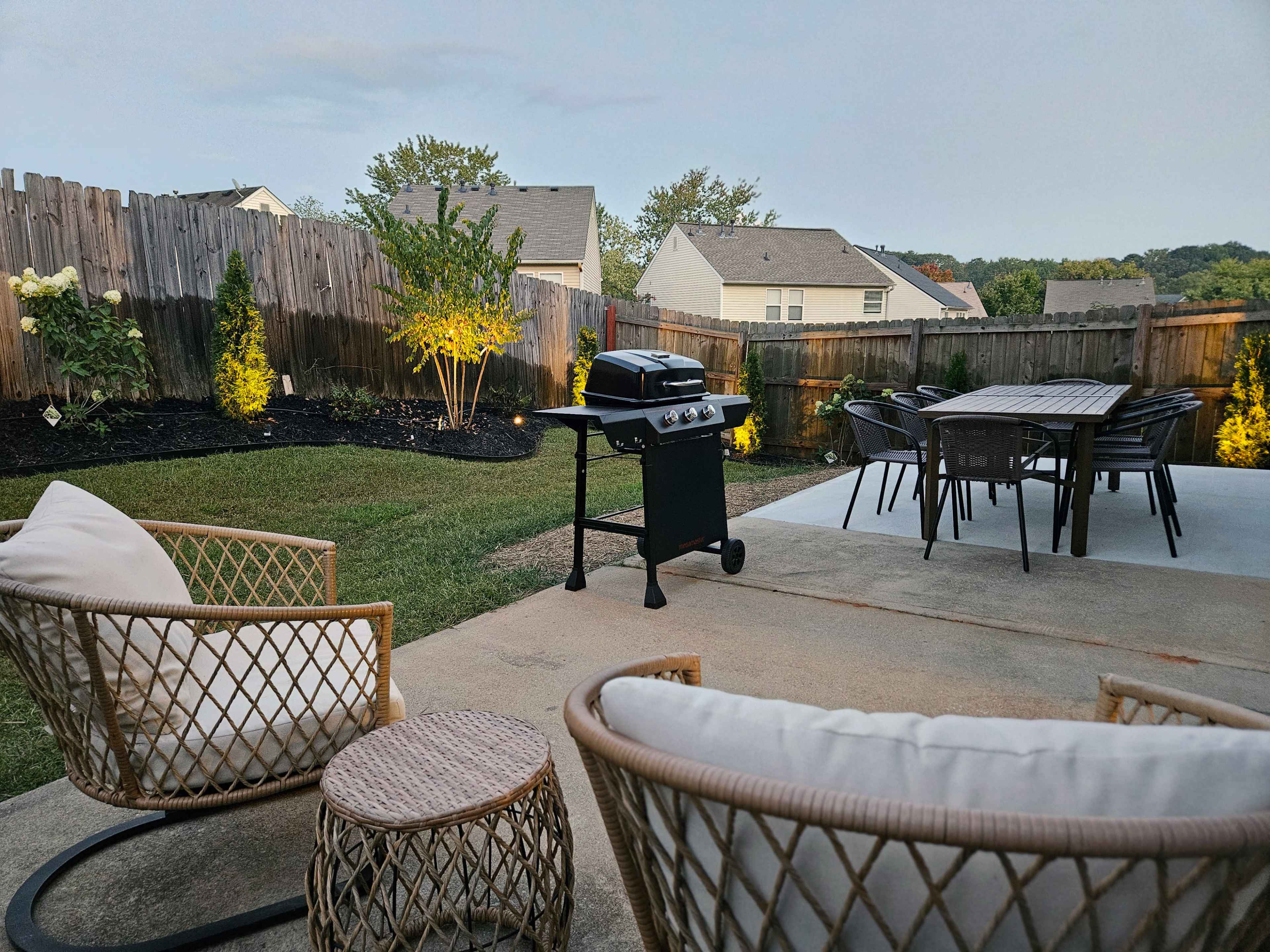 The image shows a landscaped backyard with a patio featuring a grill, outdoor seating, and decorative lighting.