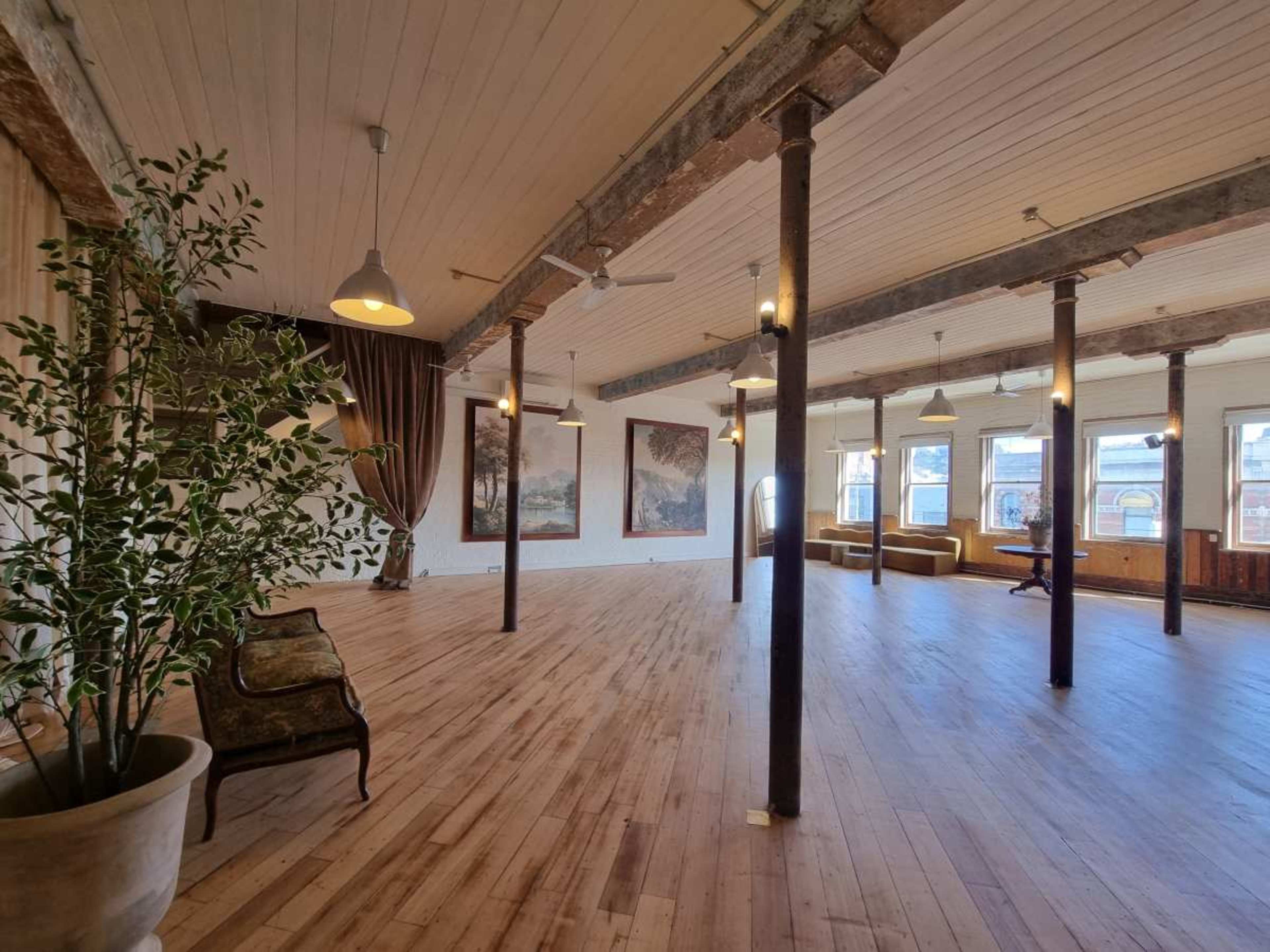 The image depicts a spacious, well-lit interior with wooden floors, tall ceilings, and decorative pillars, featuring large windows and a mix of seating areas.