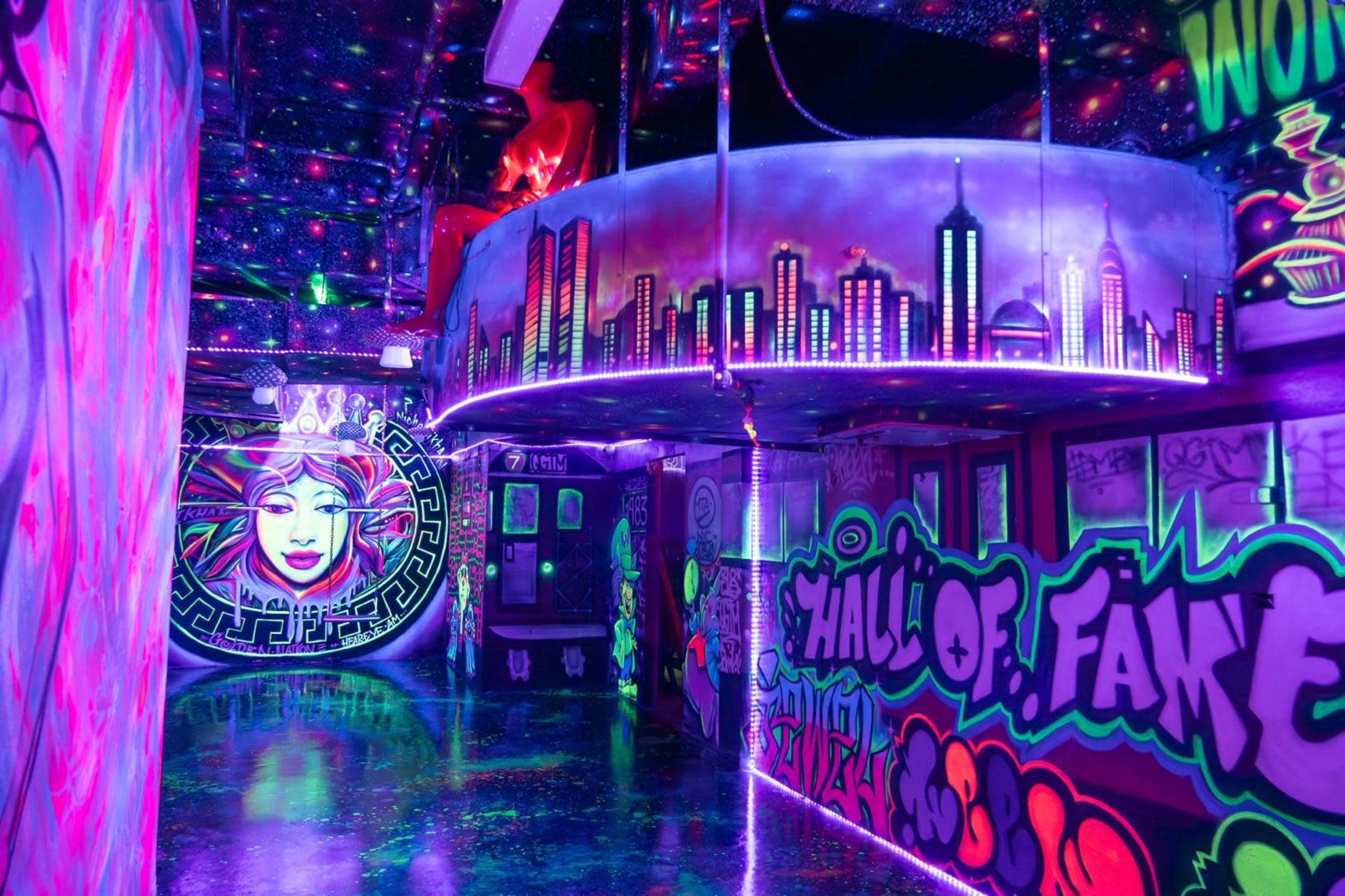 A vibrant, neon-lit hallway adorned with graffiti and illuminated by colorful lights, featuring a large mural of a face and a skyline design.