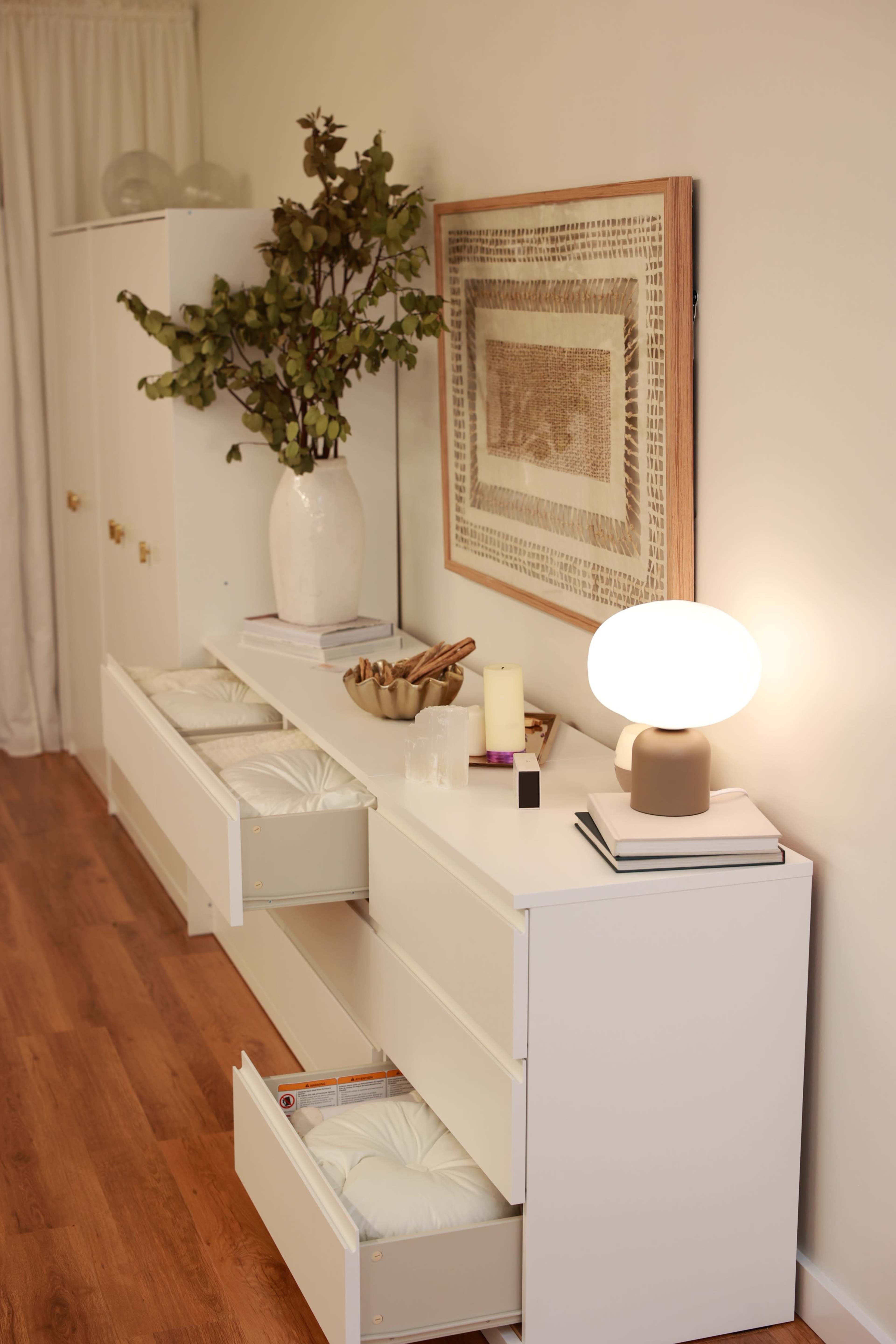 A minimalist living space features a white dresser with open drawers, a lamp, books, and a framed piece of art above, complemented by a large vase with greenery.