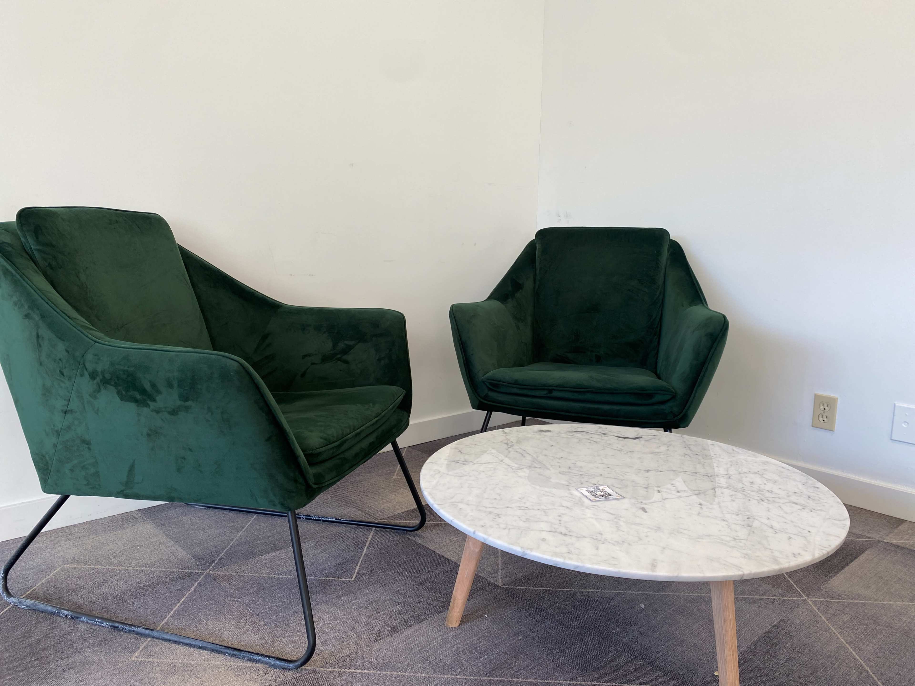Two green velvet chairs positioned next to a round marble coffee table on a gray tiled floor in a minimalist setting.