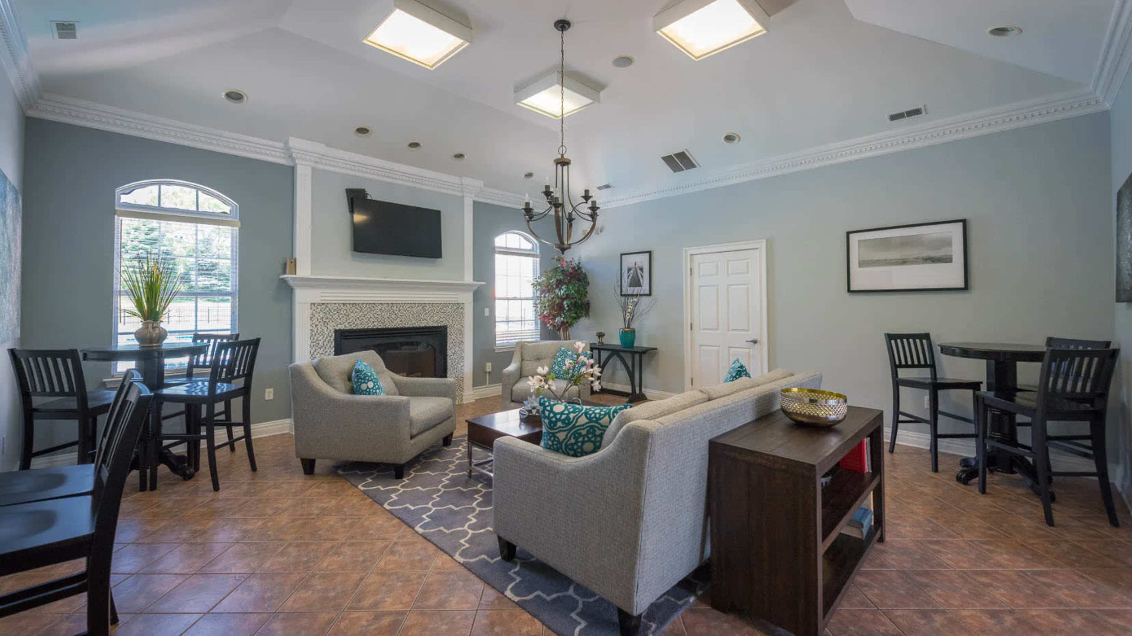 The image shows a well-lit common area featuring two gray sofas, a coffee table, and a fireplace, surrounded by dining tables and chairs.