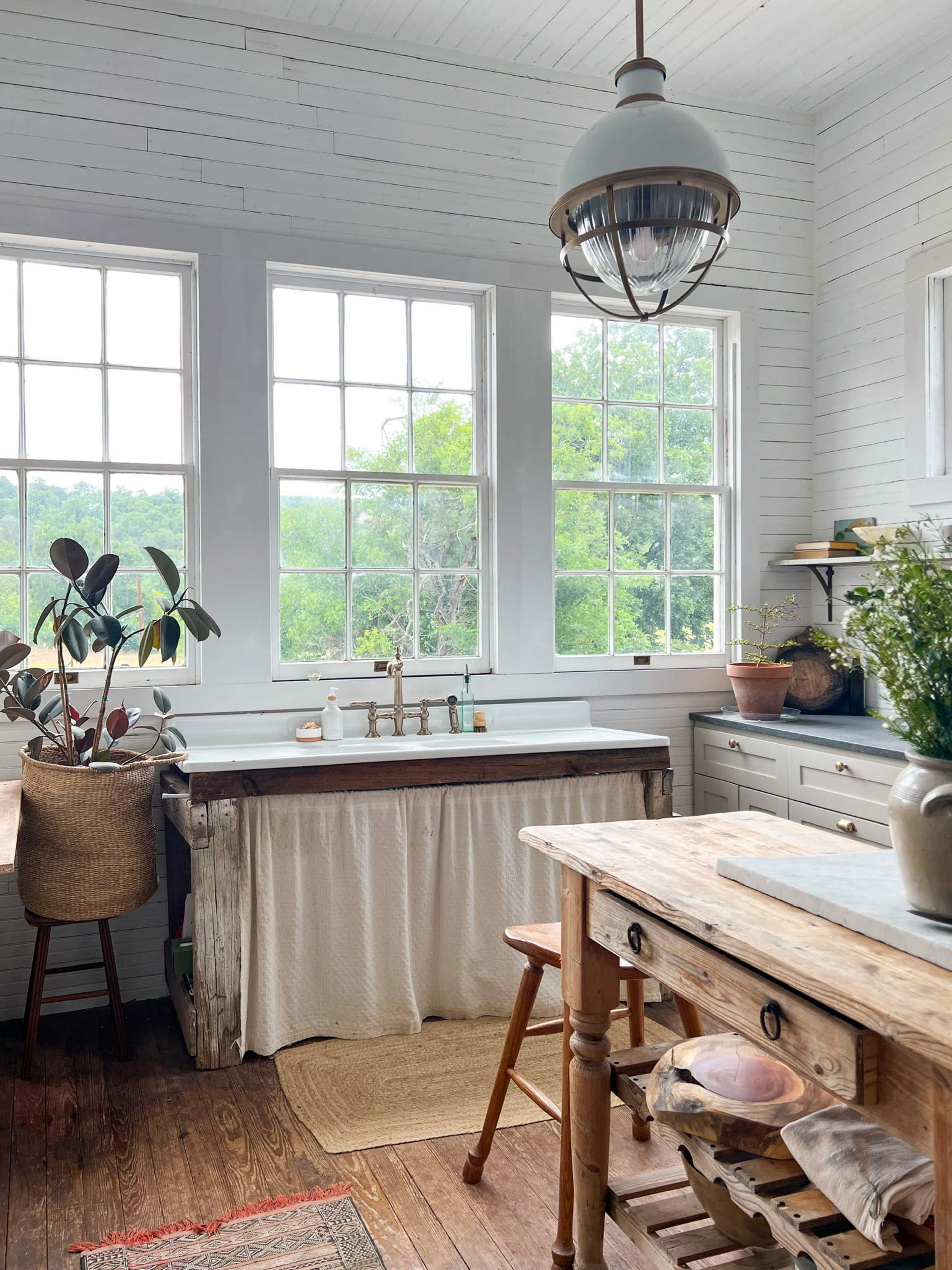 The kitchen features large windows that provide natural light, a wooden sink with a curtain underneath, and rustic furnishings including a wooden table and chair.