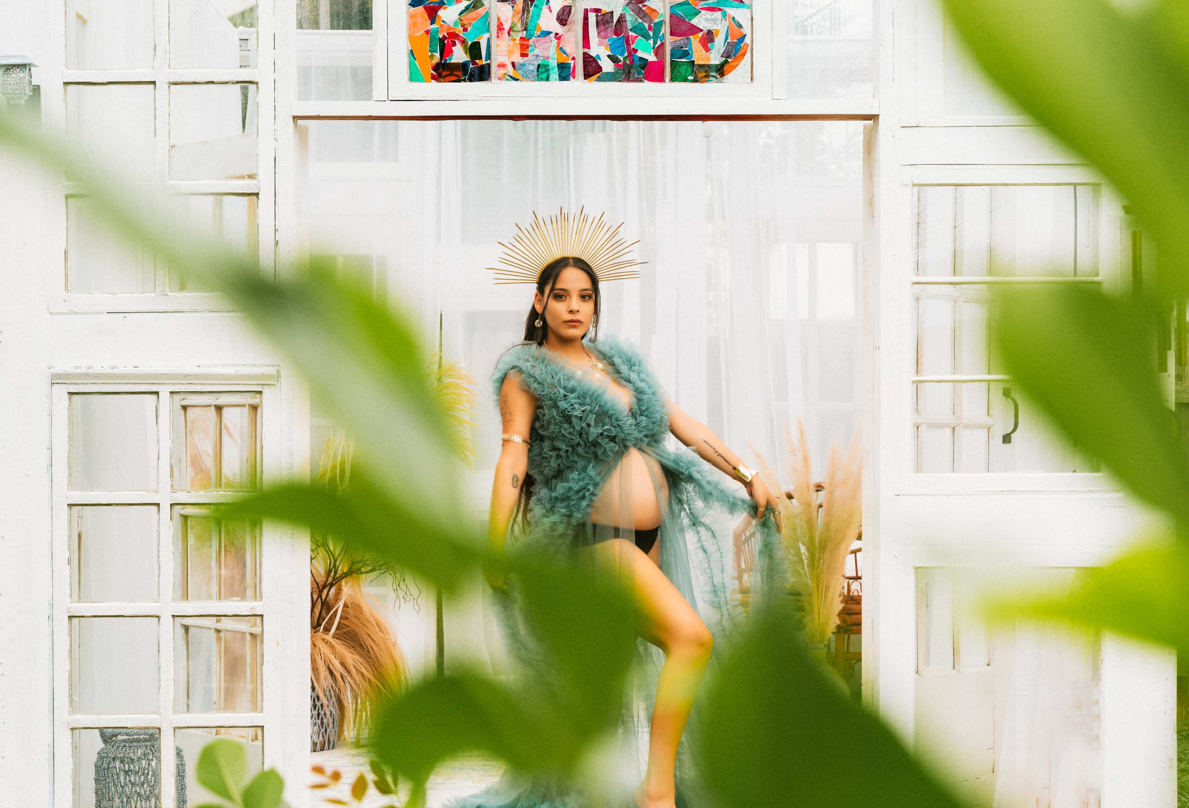 A pregnant woman poses in front of a white windowed backdrop, wearing a green feather cover and a gold crown.