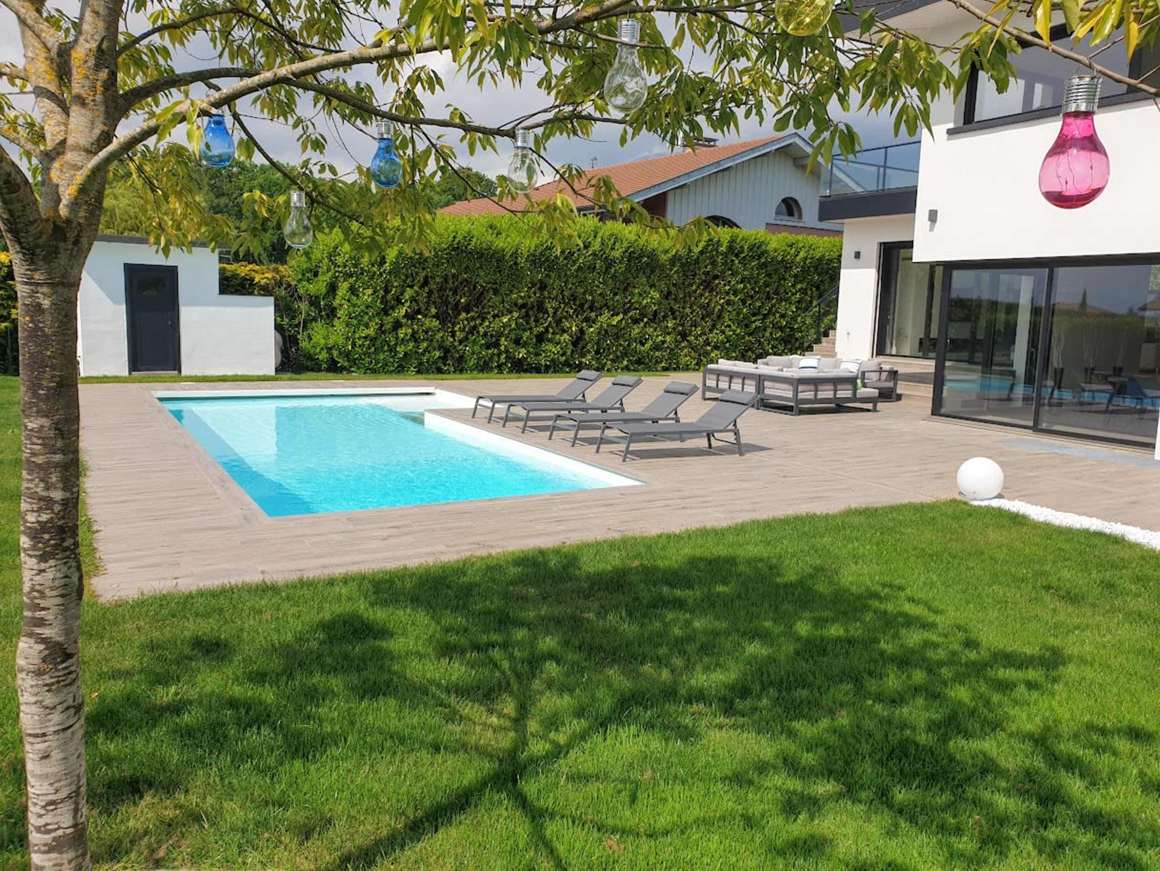 The image shows a modern backyard featuring a swimming pool, lounge chairs, and a landscaped area with grass and trees.