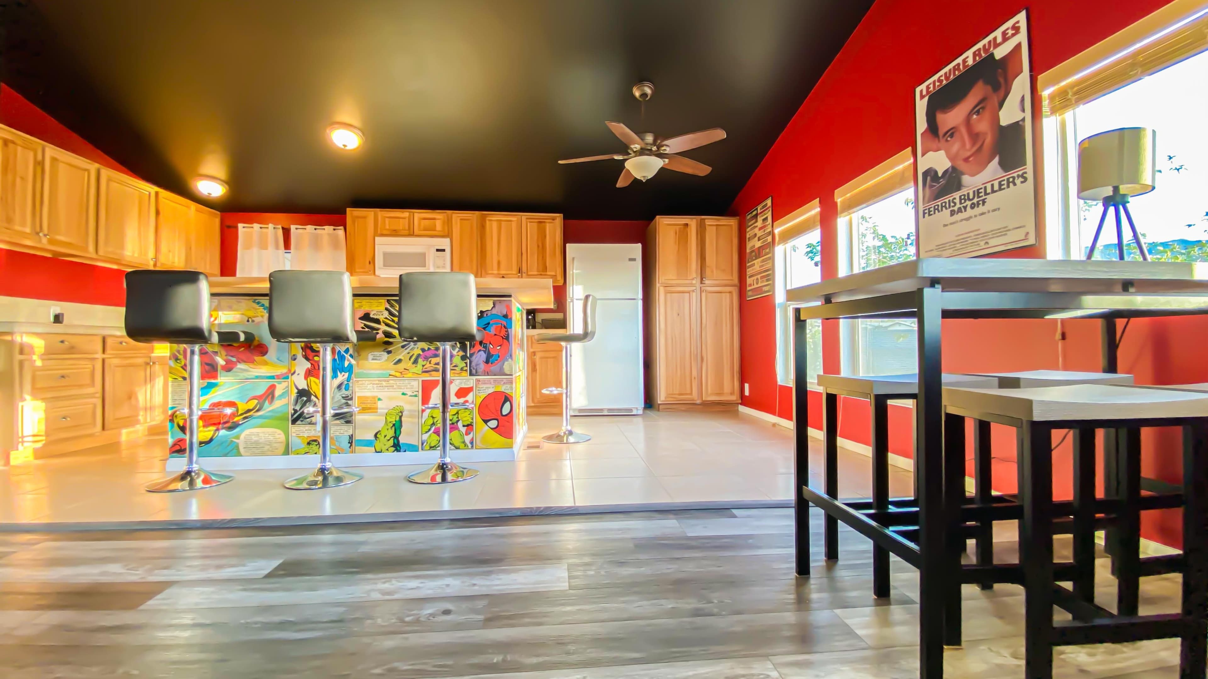 A kitchen with red walls, wooden cabinets, modern bar stools, and a table beside a window.