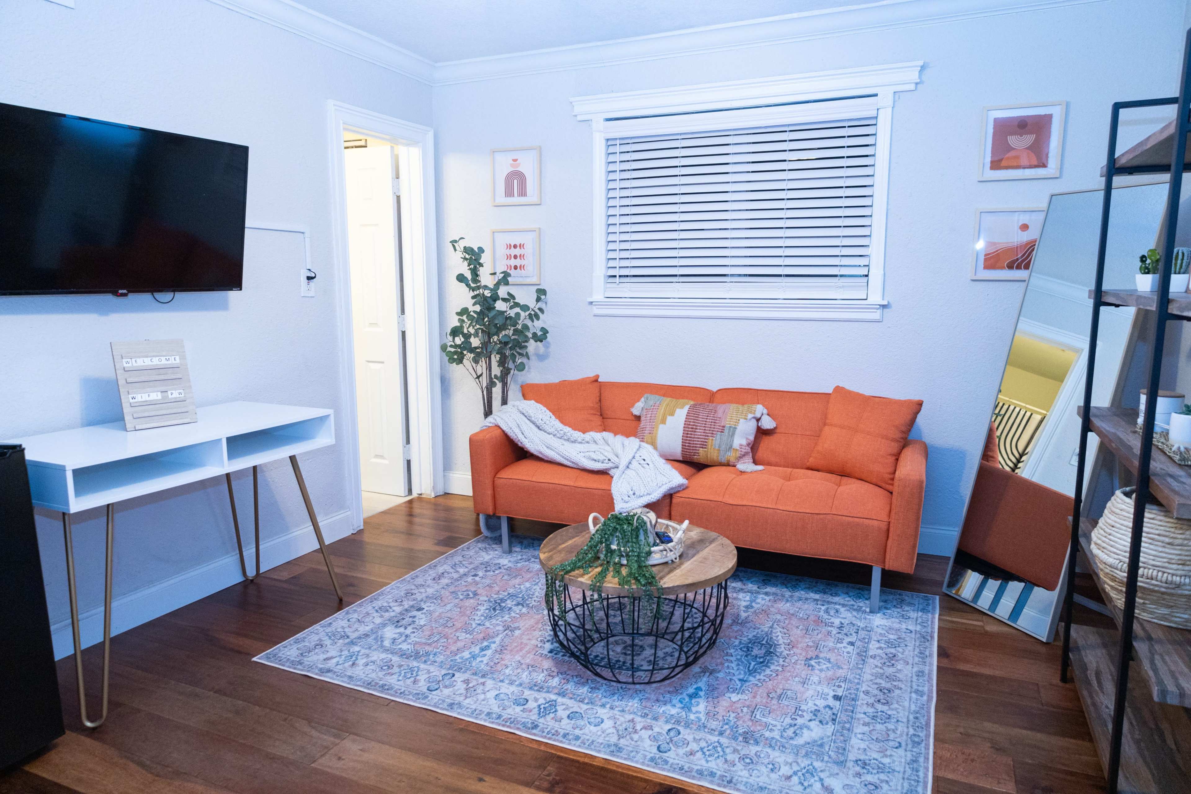 The image shows a modern living room featuring an orange sofa, a round coffee table with a decorative plant, a large mirror, and a wall-mounted TV.
