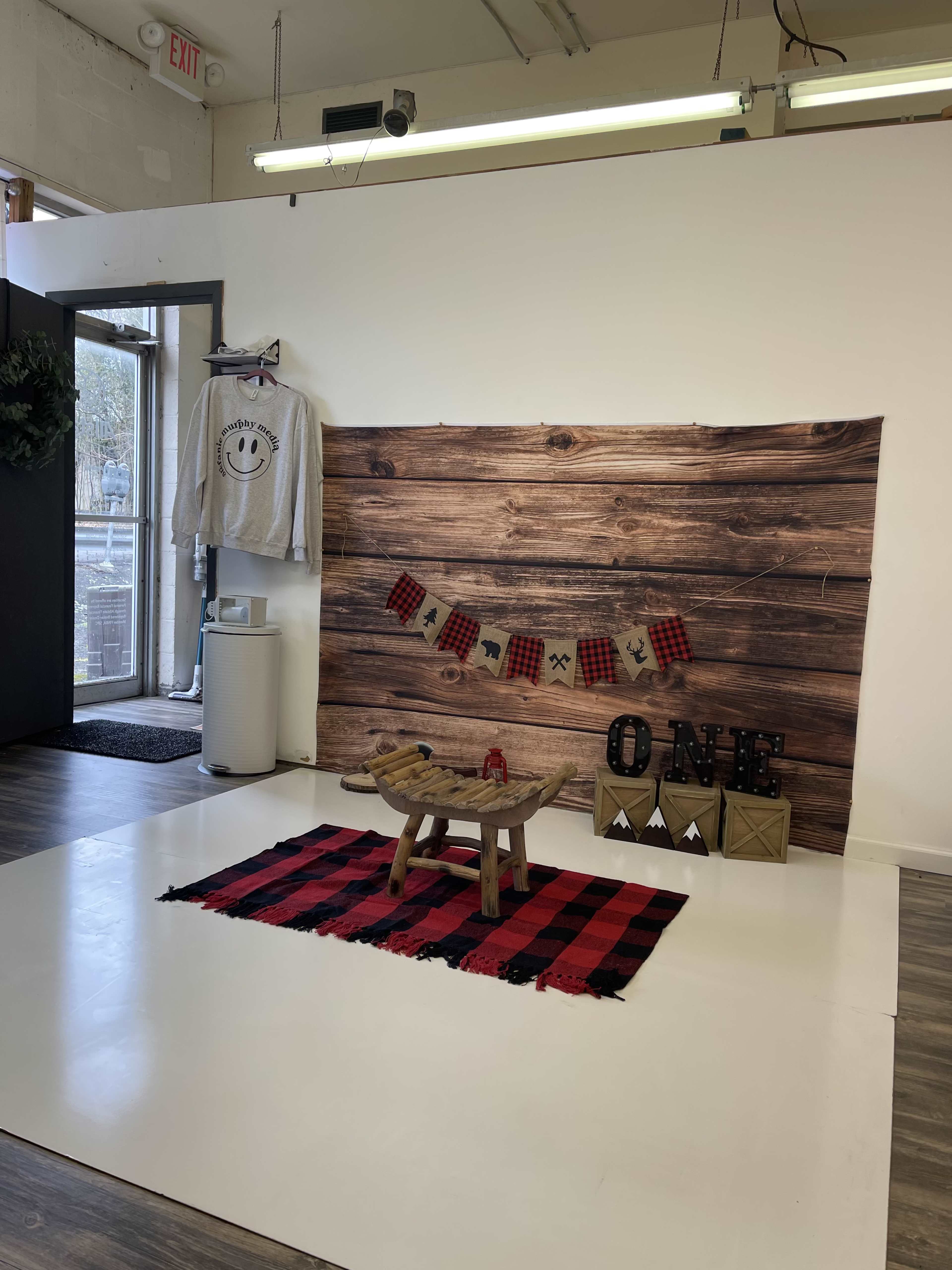 A small, styled interior space featuring a wooden backdrop, a red and black checkered rug, a wooden stool, and decorative items including a smiley sweatshirt, a banner, and letters spelling "ONE."