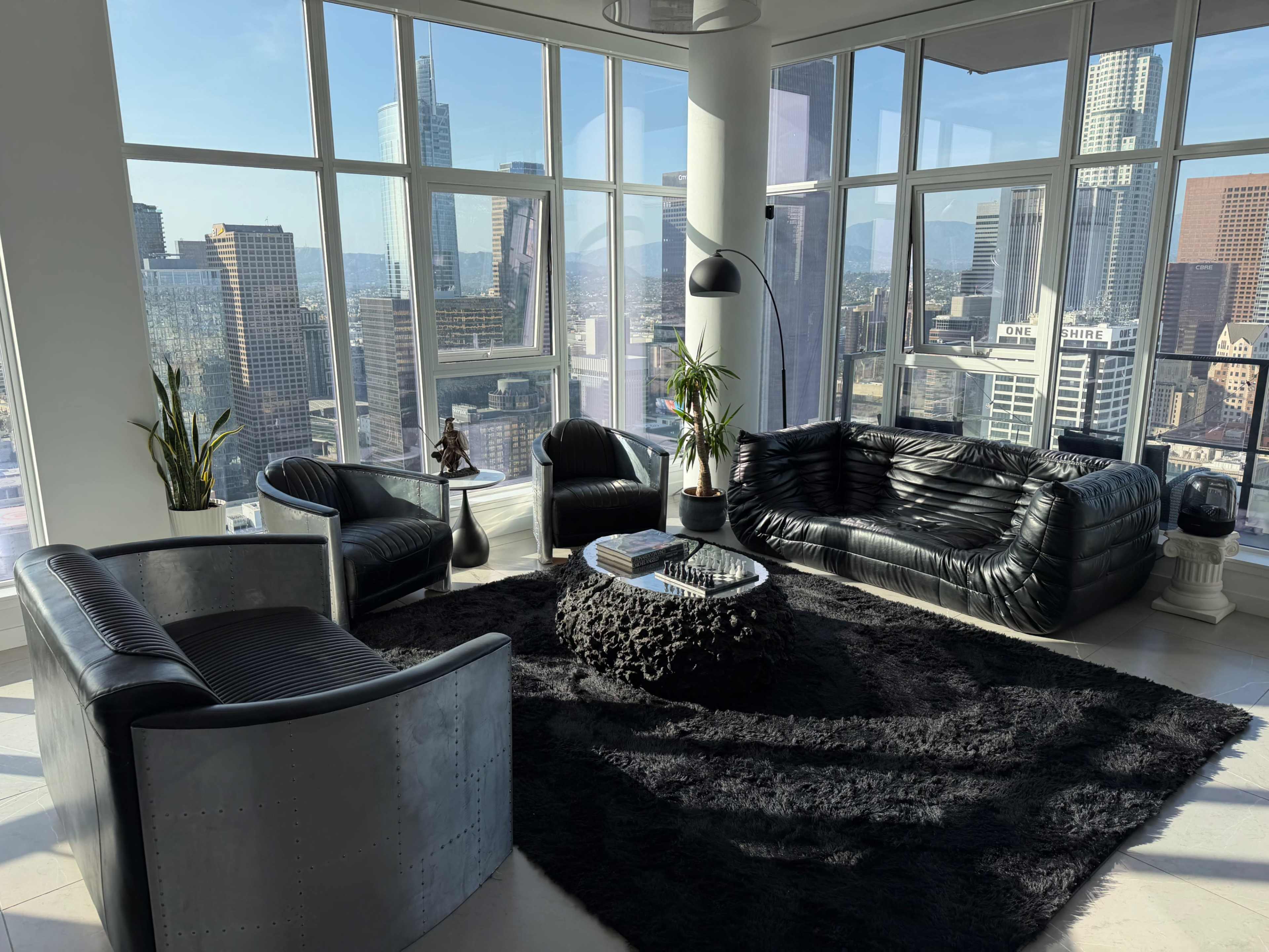 A modern living room with large windows offers a panoramic view of a city skyline, featuring black leather furniture arranged around a textured coffee table on a dark carpet.