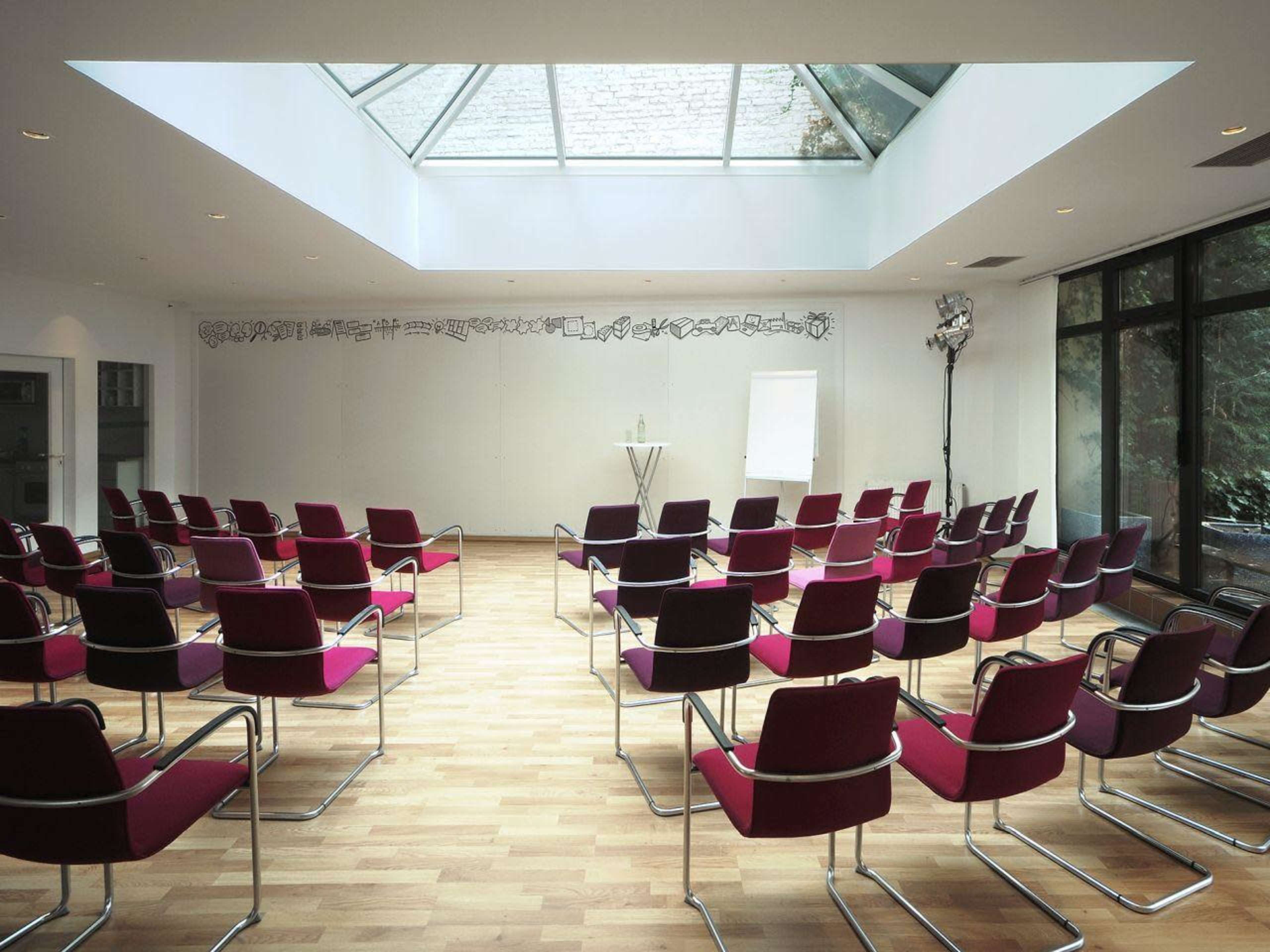 A modern conference room arranged with red and black chairs facing a whiteboard under a skylight.